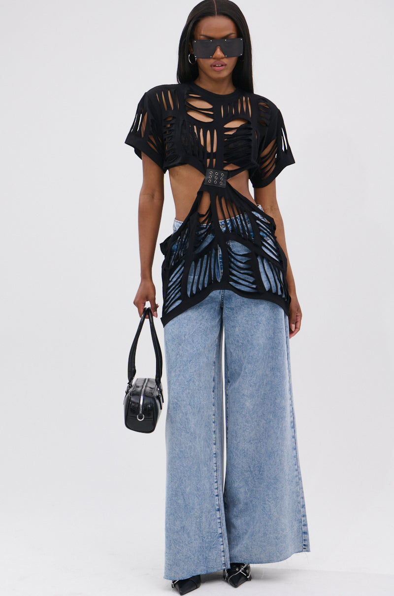 A woman wearing large sunglasses, the SHREDDED OVERSIZED SHORT SLEEVE TEE with bold patterns, light blue wide-leg jeans, black pointed shoes, and a small black handbag stands against a plain white background.