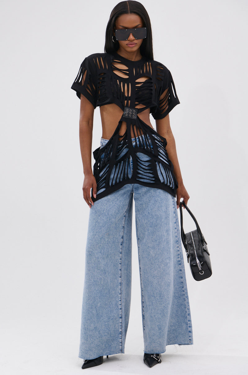 Wearing the SHREDDED OVERSIZED SHORT SLEEVE TEE, wide-leg light blue jeans with grommet belt, pointed black shoes, and sunglasses, they hold a black handbag while standing against a plain white background.