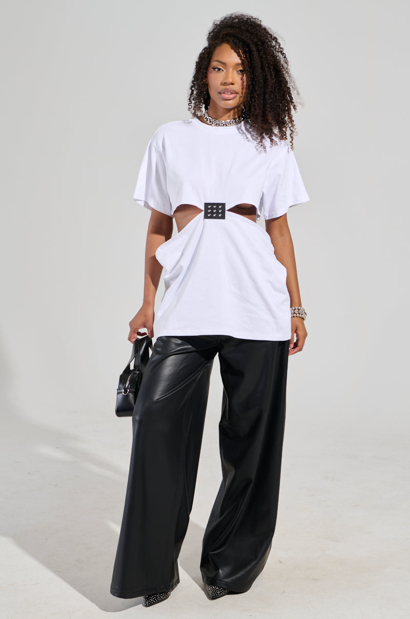 A person showcases a chic look with a unique design, wearing the GIRL BOSS MOMENTS CUT OUT T-SHIRT in white, along with wide-leg black leather pants. They hold a small black handbag, their curly hair styled naturally against a neutral background.