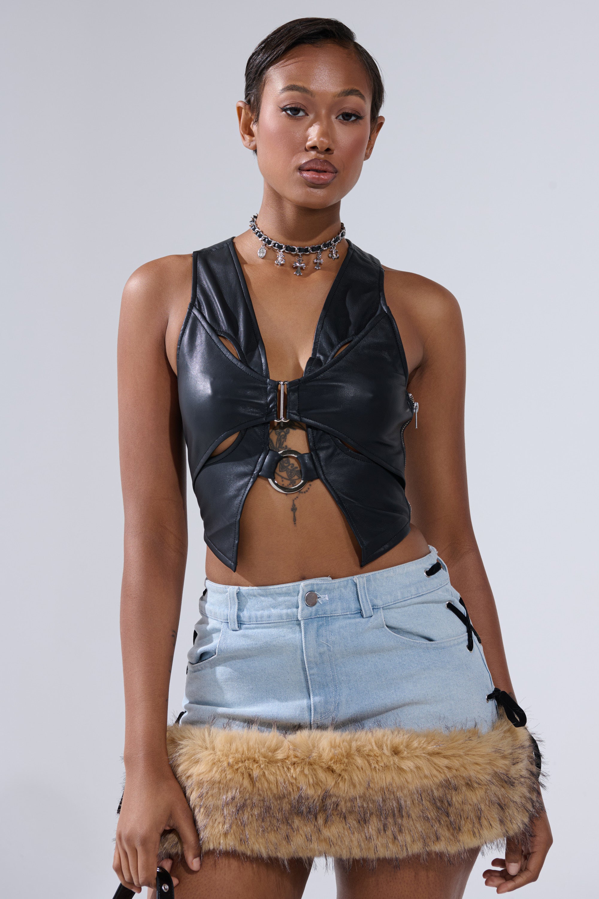 A woman stands confidently against a plain background wearing the FLASHY QUEEN BRALETTE, a black cutout leather top styled with a choker and a light denim mini skirt featuring tan faux fur trim at the hem.