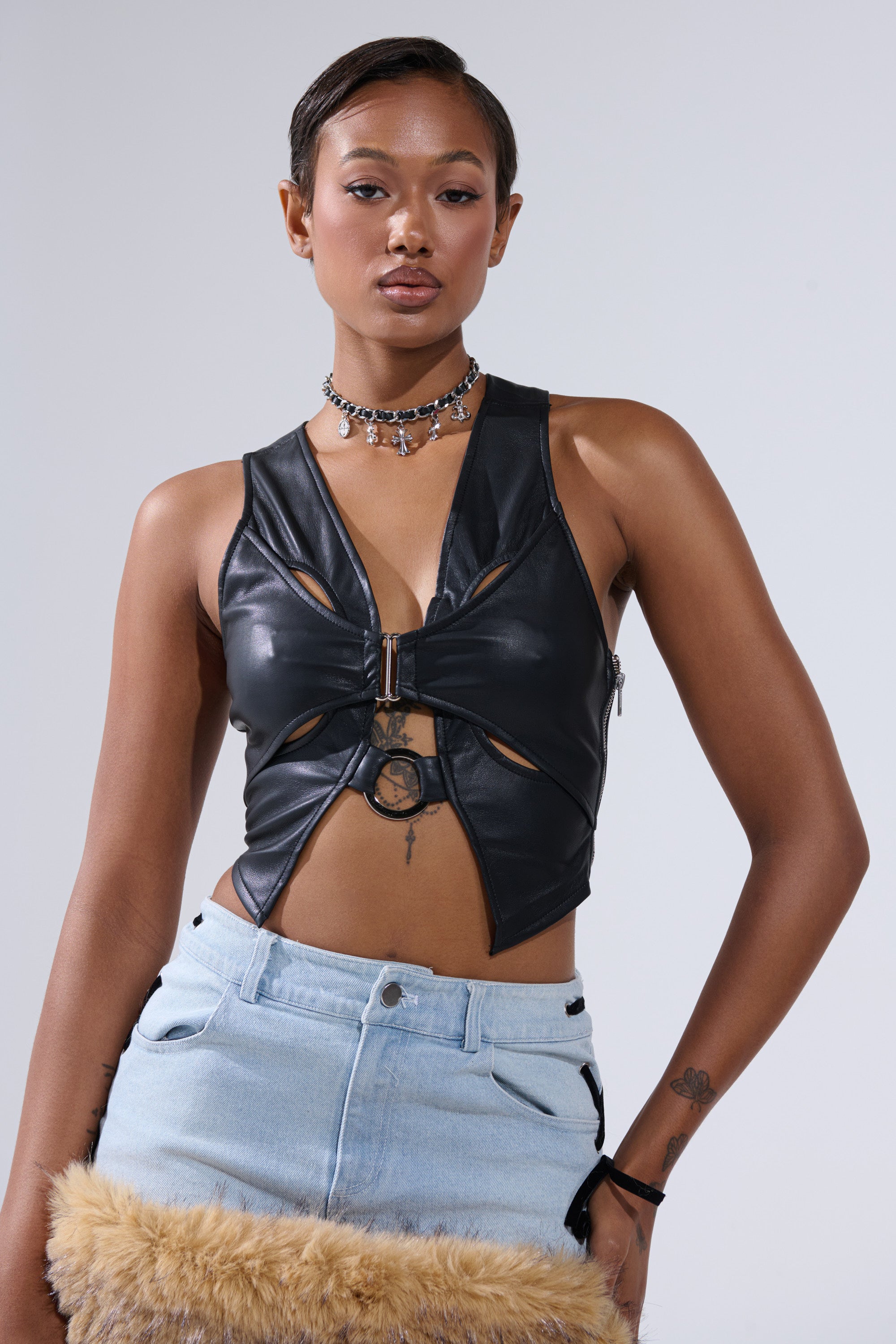 A woman stands against a plain background wearing the FLASHY QUEEN BRALETTE, a black cutout leather top, paired with a light blue denim zipper skirt, a layered choker necklace, and holding a furry tan accessory.