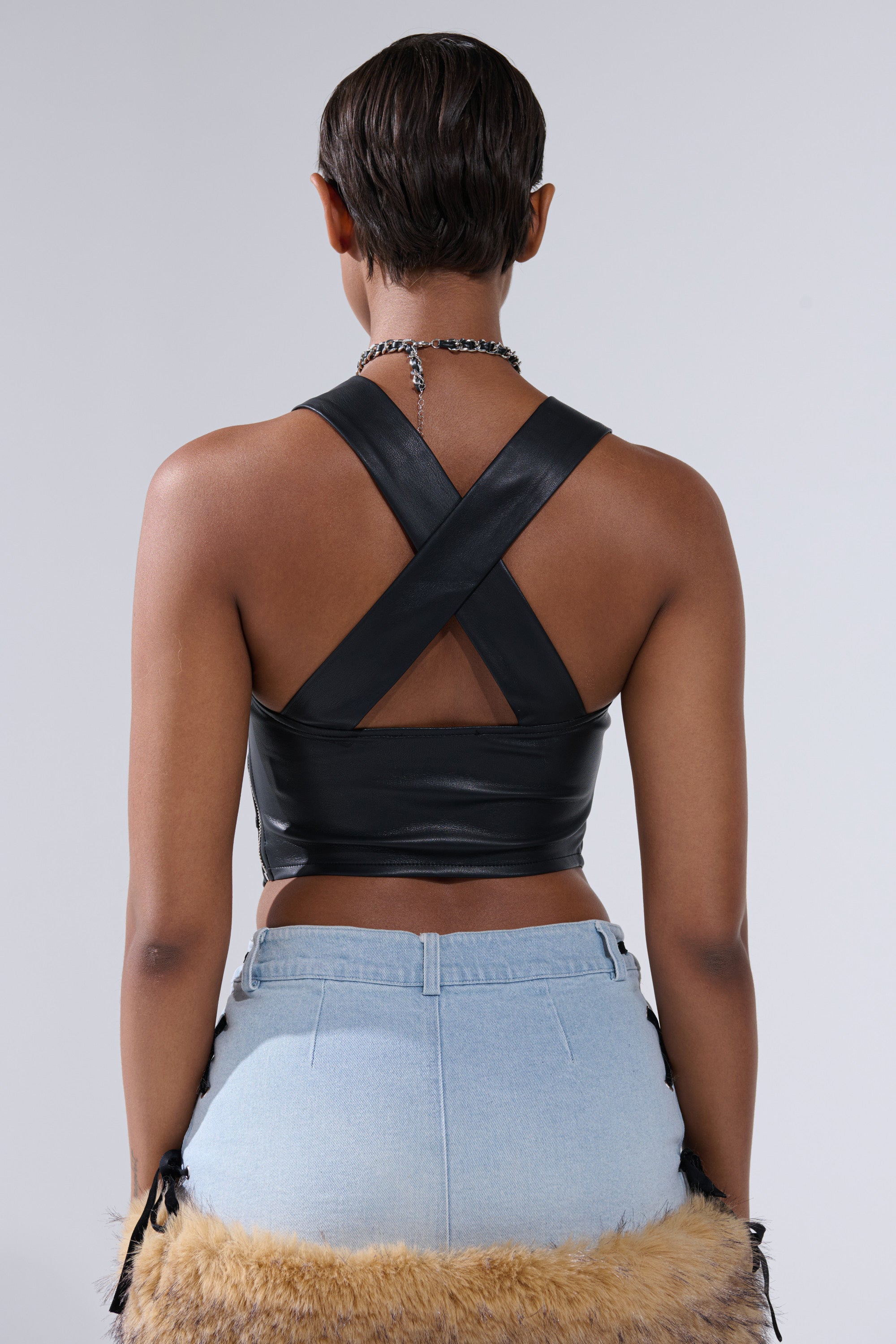 A person faces away from the camera, wearing the FLASHY QUEEN BRALETTE— a black faux leather cross-back crop top—paired with light blue high-waisted jeans, a necklace, and a faux fur accent on the lower outfit against a plain gray background.