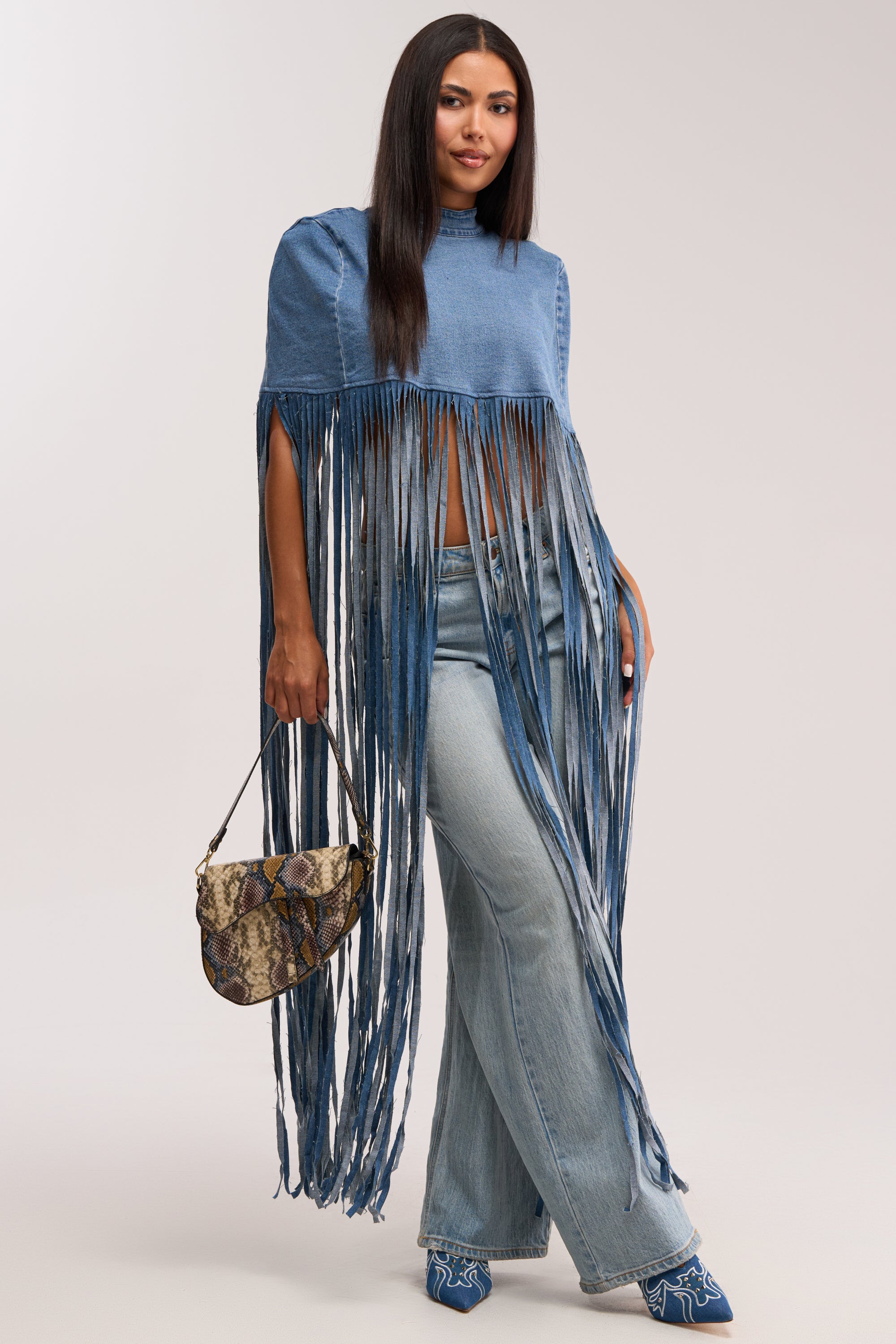 A woman models the DENIM DREAMS BLOUSE with wide-leg jeans and embroidered blue shoes, holding a small snakeskin-patterned handbag. Her long dark hair and the beige background highlight her chic western style.