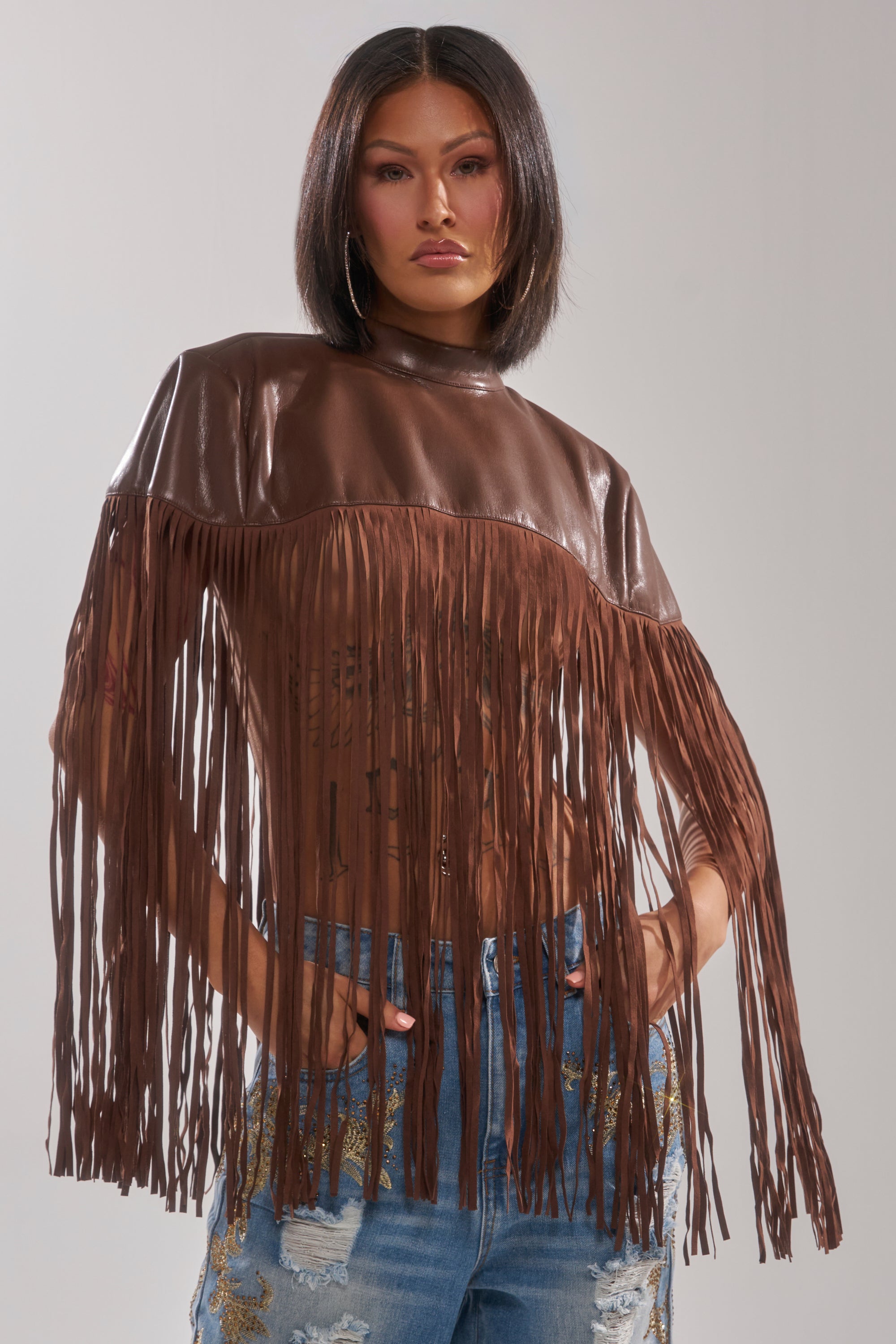 A woman with straight dark hair models the FRINGE FOR DAYS FAUX LEATHER TOP, paired with ripped blue jeans featuring gold embroidery—an ideal layering piece for festivals. She poses with hands in her pockets against a simple light backdrop.