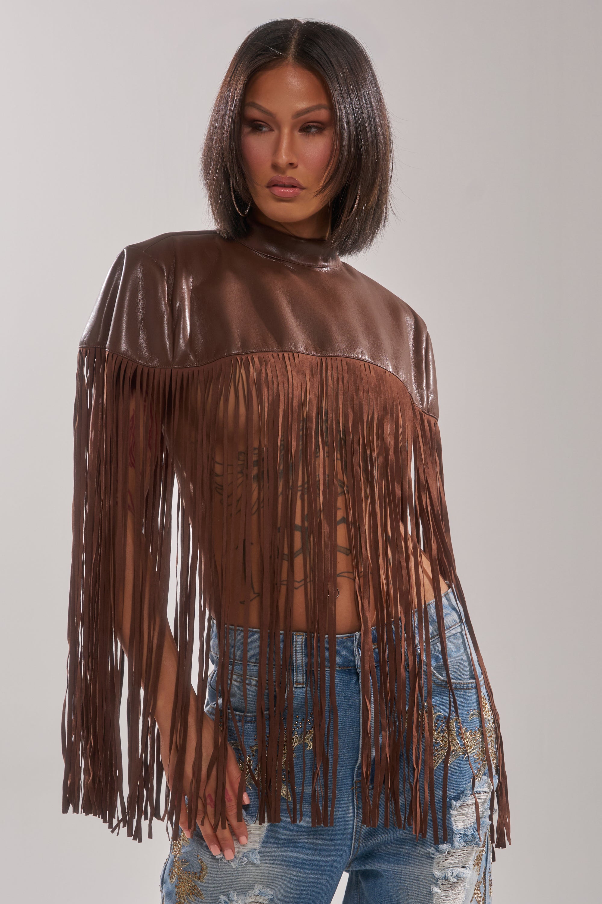 A woman with a bob poses against a plain background wearing the FRINGE FOR DAYS FAUX LEATHER TOP, styled with distressed blue jeans featuring gold embroidery.