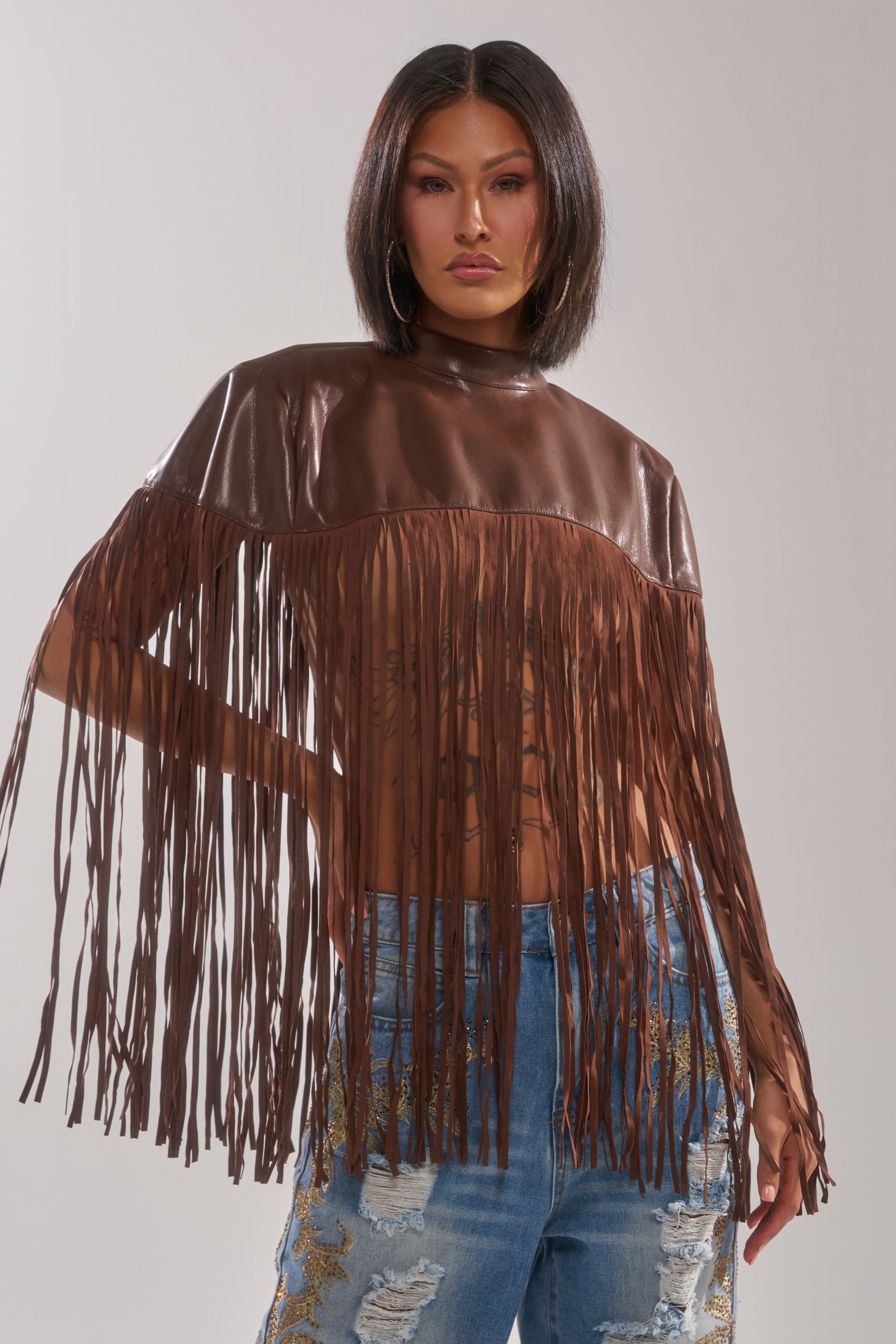 A woman with straight dark hair wears the FRINGE FOR DAYS FAUX LEATHER TOP in shiny brown and distressed blue jeans with gold embroidery, standing confidently against a plain light background.