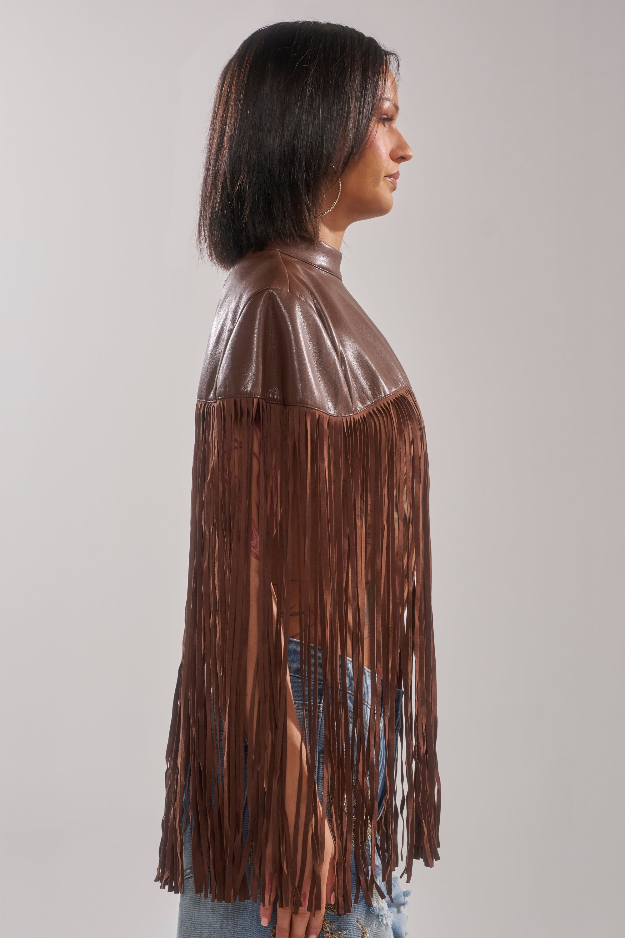 A woman stands in profile wearing the FRINGE FOR DAYS FAUX LEATHER TOP with long brown fringe from shoulders to wrists, paired with light blue jeans—a perfect festival outfit set against a plain background.