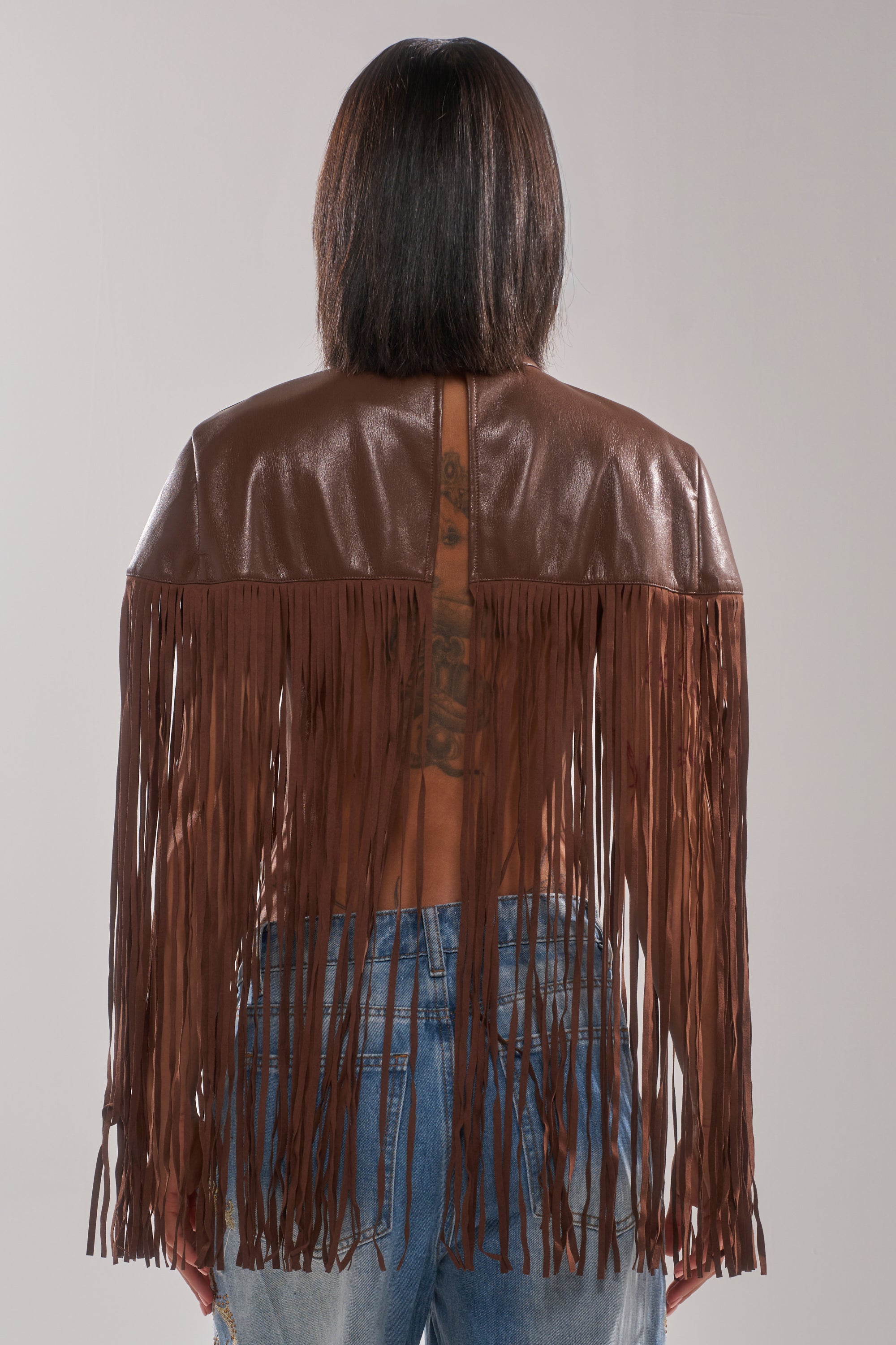 A person with straight, shoulder-length hair models the FRINGE FOR DAYS FAUX LEATHER TOP and blue jeans from behind, showing a tattoo along their spine beneath the long fringe of this Western-inspired layering piece.