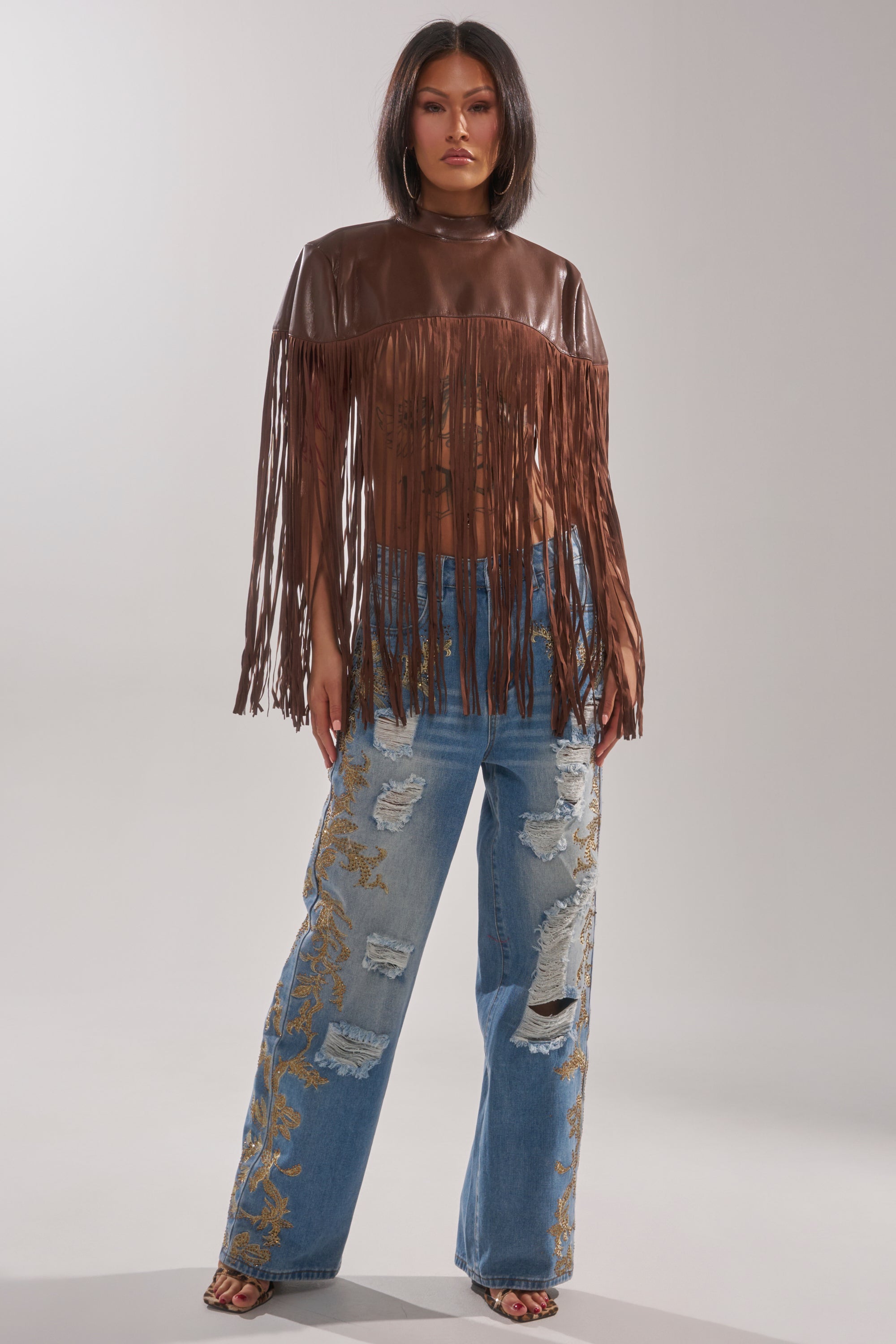 A woman models the FRINGE FOR DAYS FAUX LEATHER TOP, a brown festival layering piece with fringe, styled with wide-leg ripped jeans featuring gold embroidery against a plain light background.