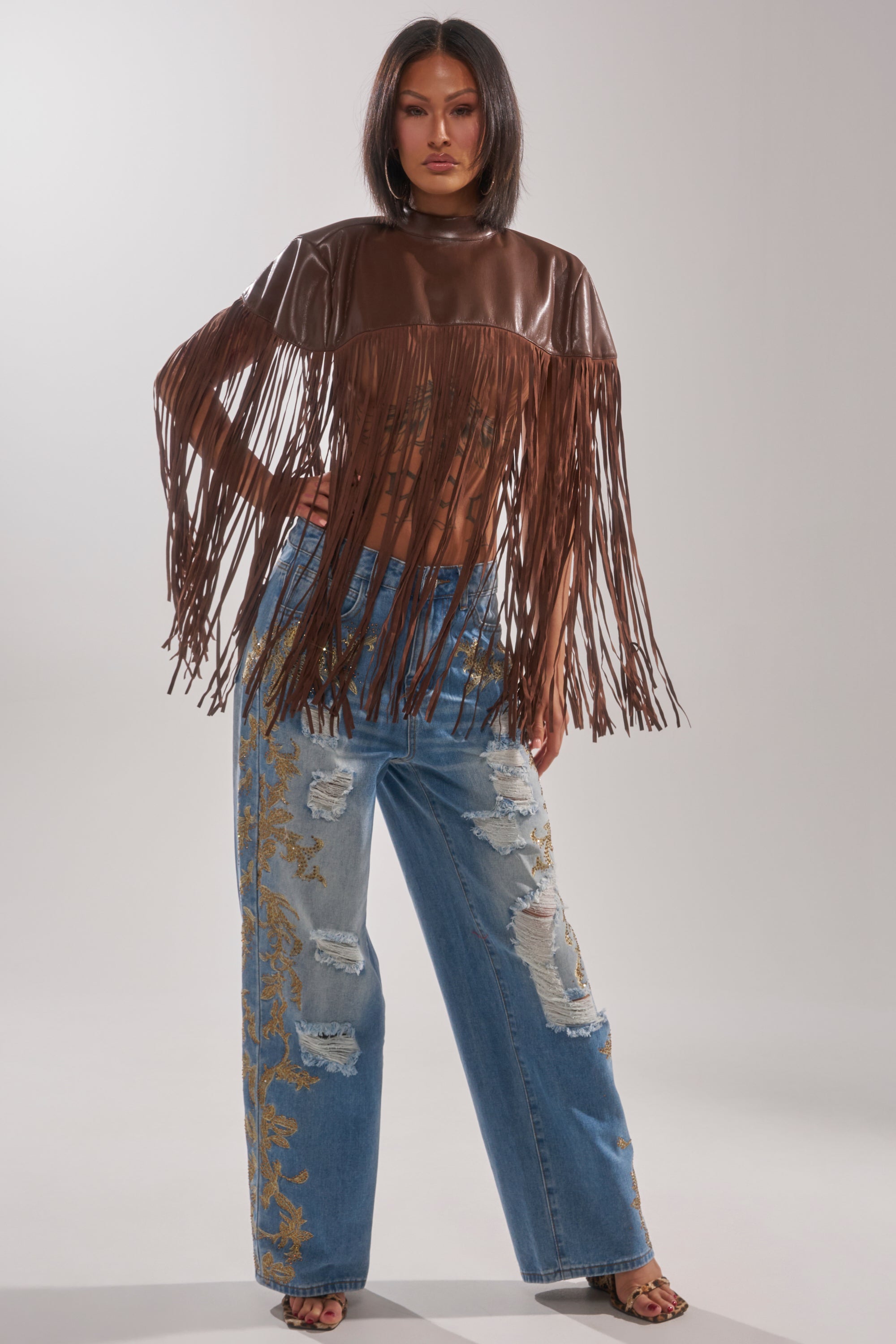 A woman models the FRINGE FOR DAYS FAUX LEATHER TOP, paired with distressed blue jeans featuring gold floral embroidery and open-toe heels, striking a confident pose against a plain light background for a chic festival look.