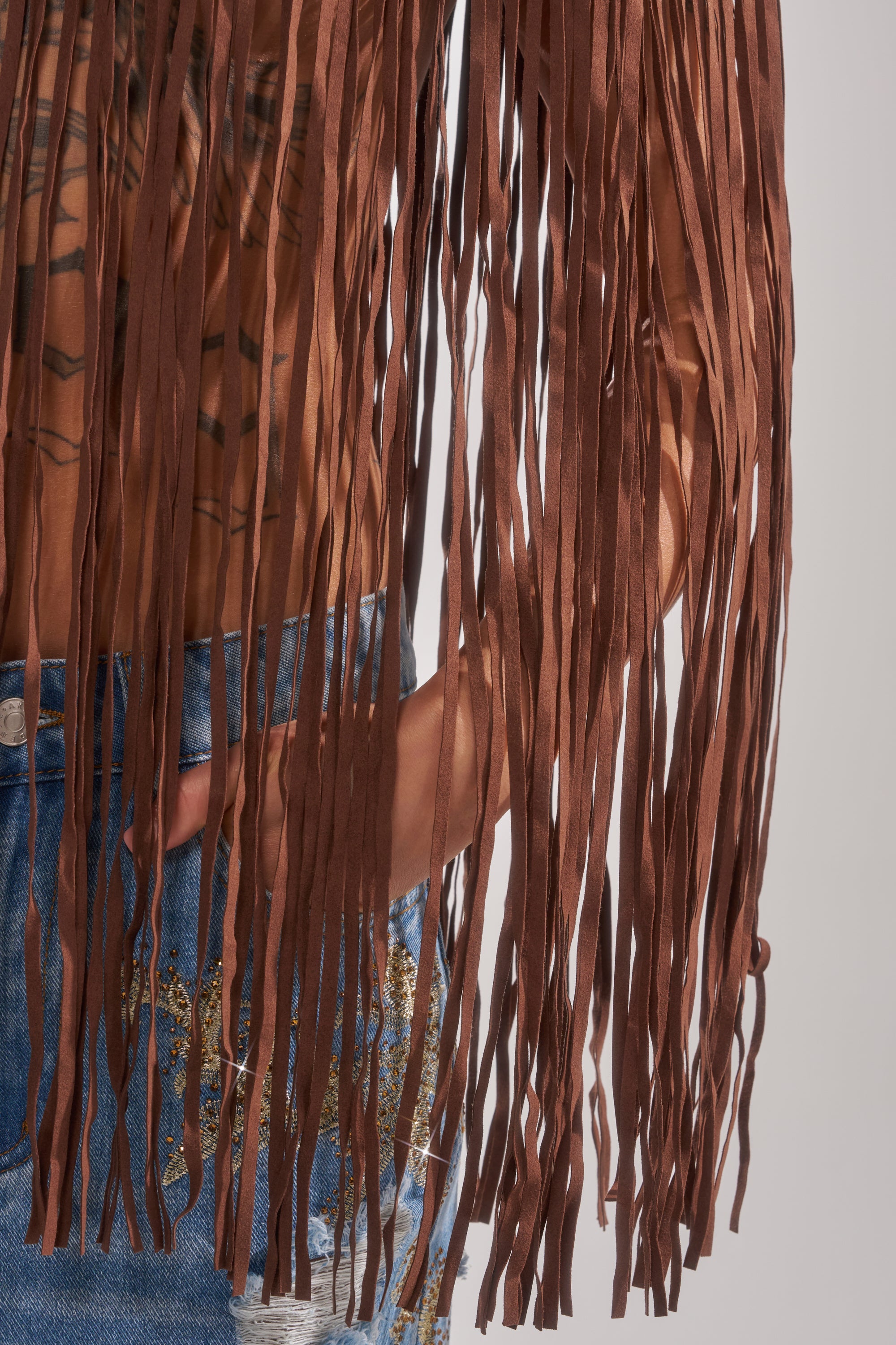 Wearing the FRINGE FOR DAYS FAUX LEATHER TOP with long brown fringe and blue jeans with gold details, their hand slips into a pocket as the fringe drapes elegantly—a perfect festival-ready look.