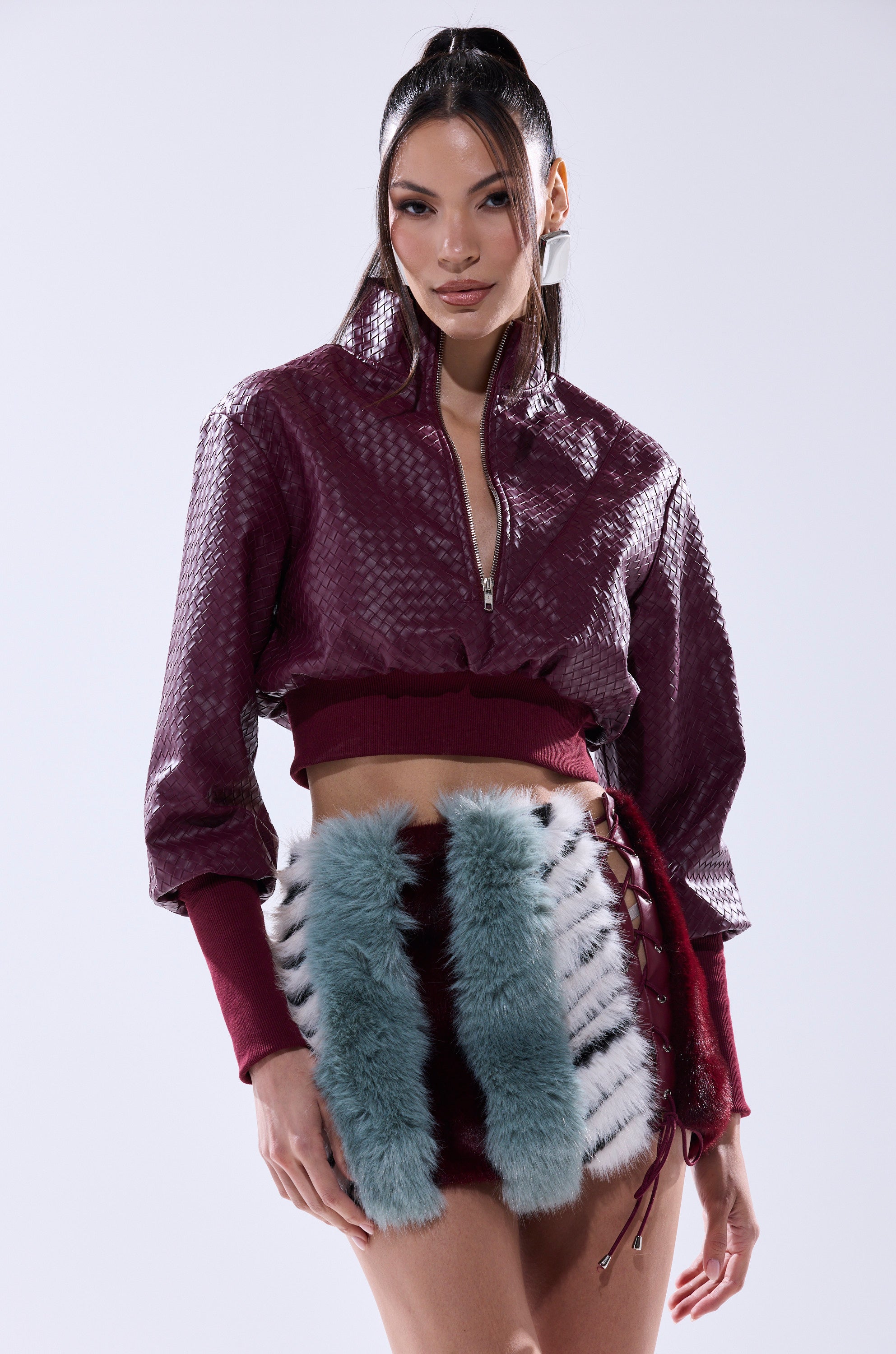 A woman poses in a maroon textured cropped jacket and mini skirt with blue and white faux fur panels, styled with the VENETTA FAUX LEATHER QUARTER ZIP SWEATSHIRT, against a plain light background.