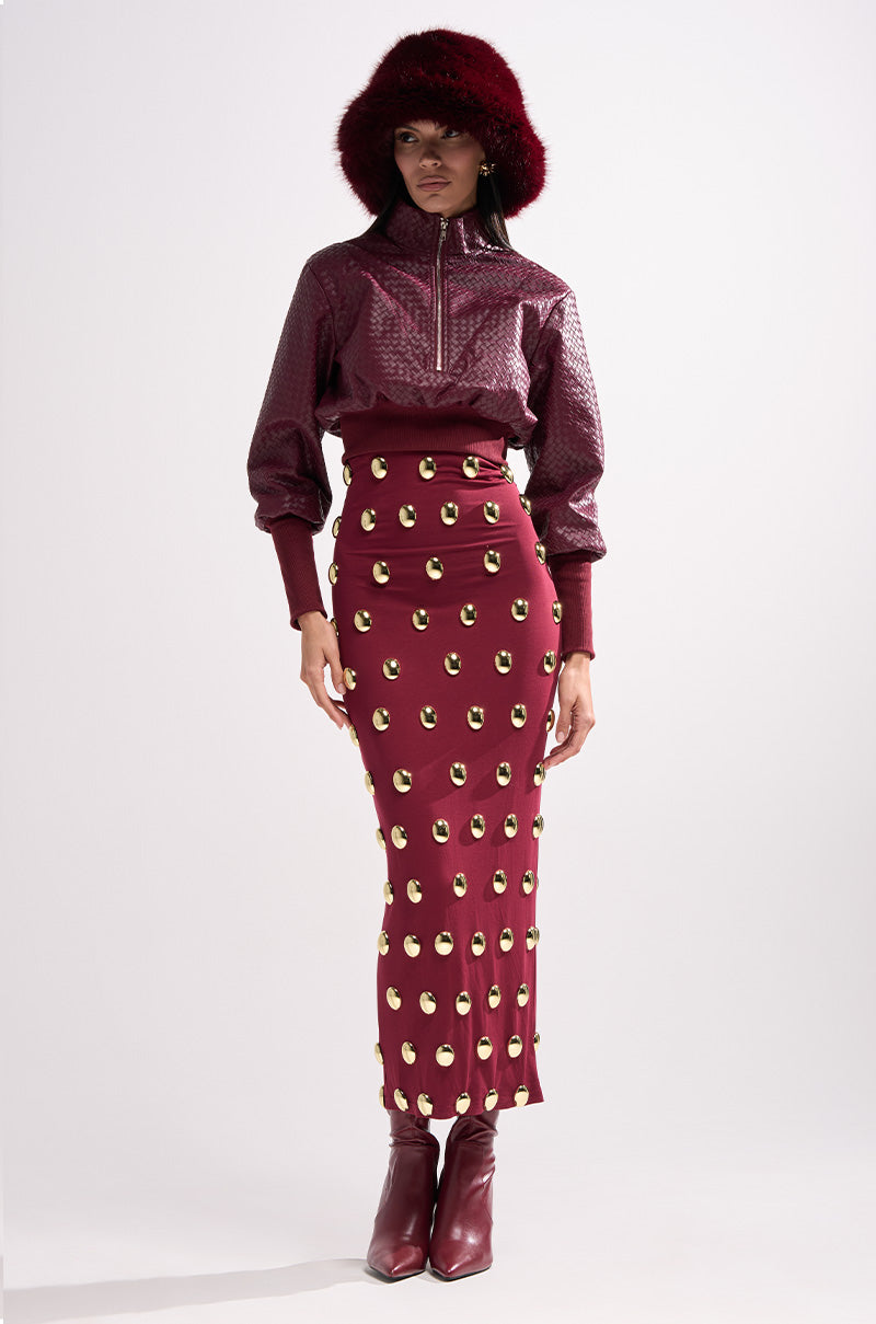 A woman in a dark red fur hat, the VENETTA FAUX LEATHER QUARTER ZIP SWEATSHIRT, a high-waisted burgundy skirt with gold details, and matching burgundy boots stands against a plain white background.