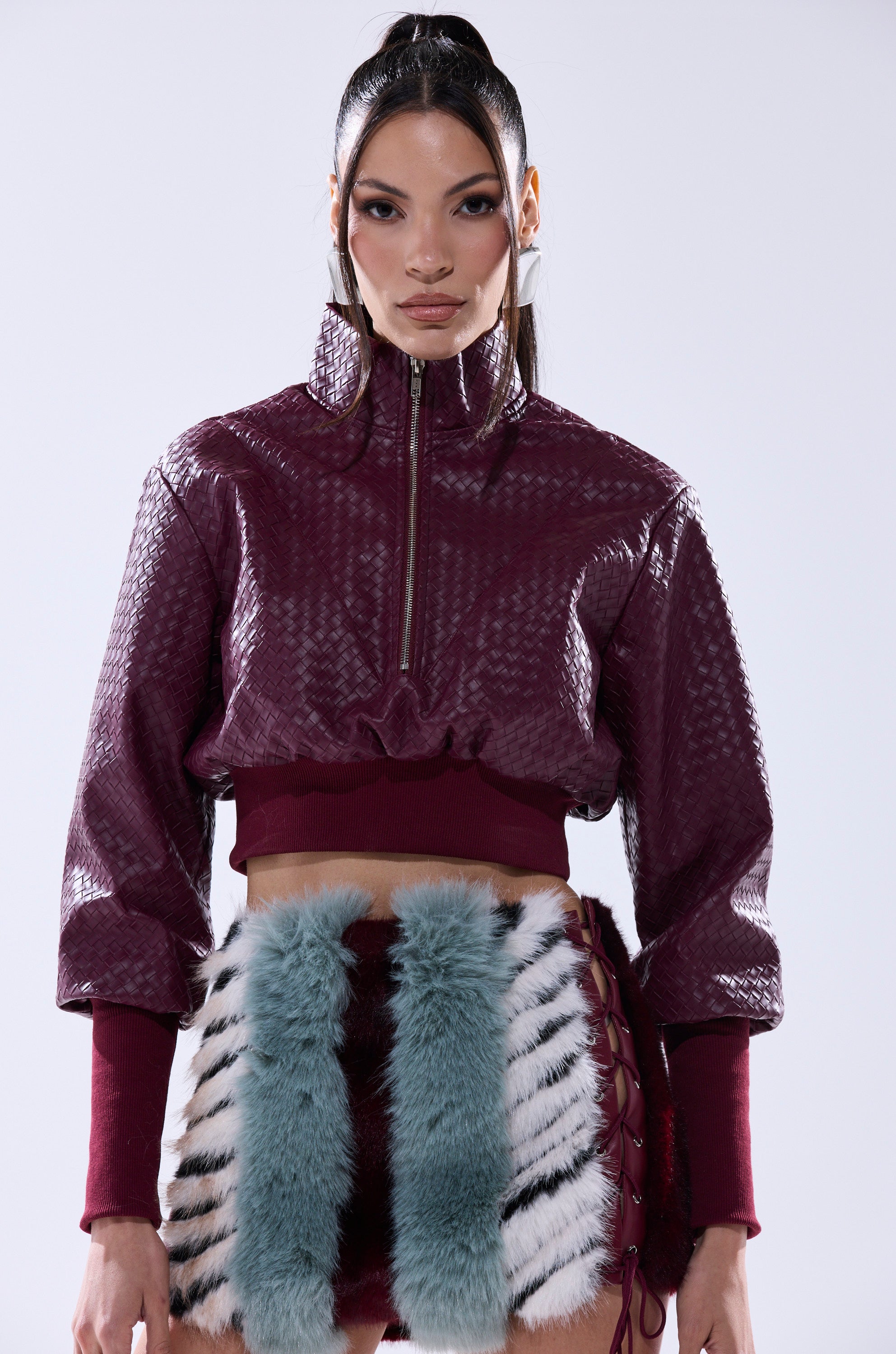 A woman poses against a plain light background in the VENETTA FAUX LEATHER QUARTER ZIP SWEATSHIRT, maroon with textured detailing, paired with a skirt featuring fluffy teal and white vertical stripes.