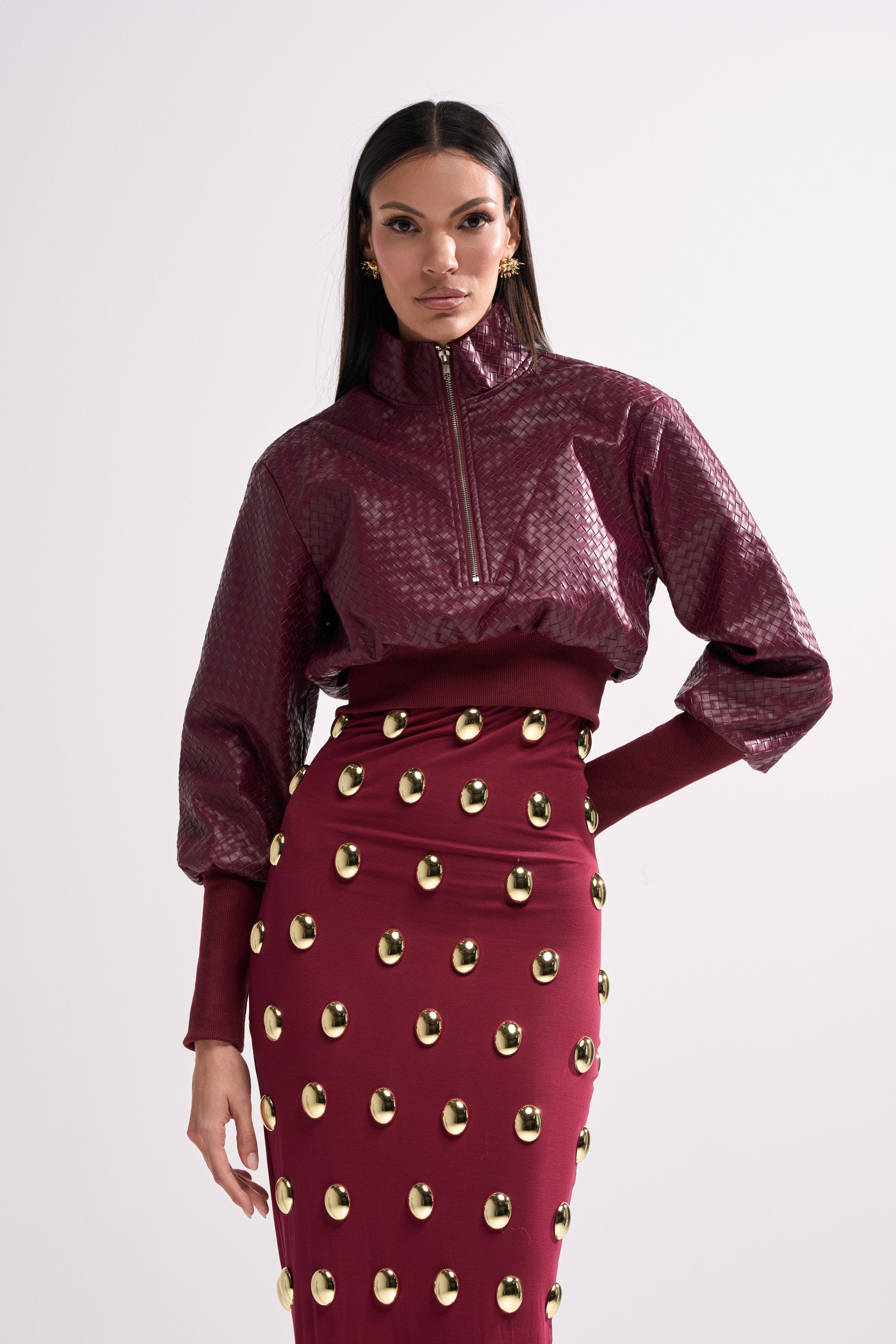 A woman models the VENETTA FAUX LEATHER QUARTER ZIP SWEATSHIRT with a matching high-waisted skirt adorned with gold studs, both featuring a shiny, textured finish, against a plain white background.