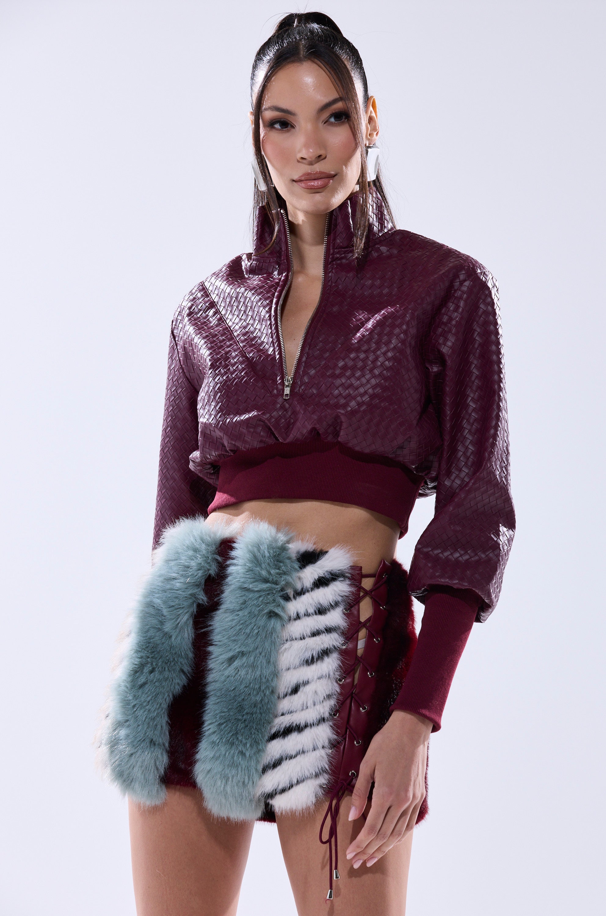 A woman poses in the VENETTA FAUX LEATHER QUARTER ZIP SWEATSHIRT, a textured burgundy cropped top, paired with high-waisted shorts with blue and white faux fur panels and lace-up sides, against a plain light background.