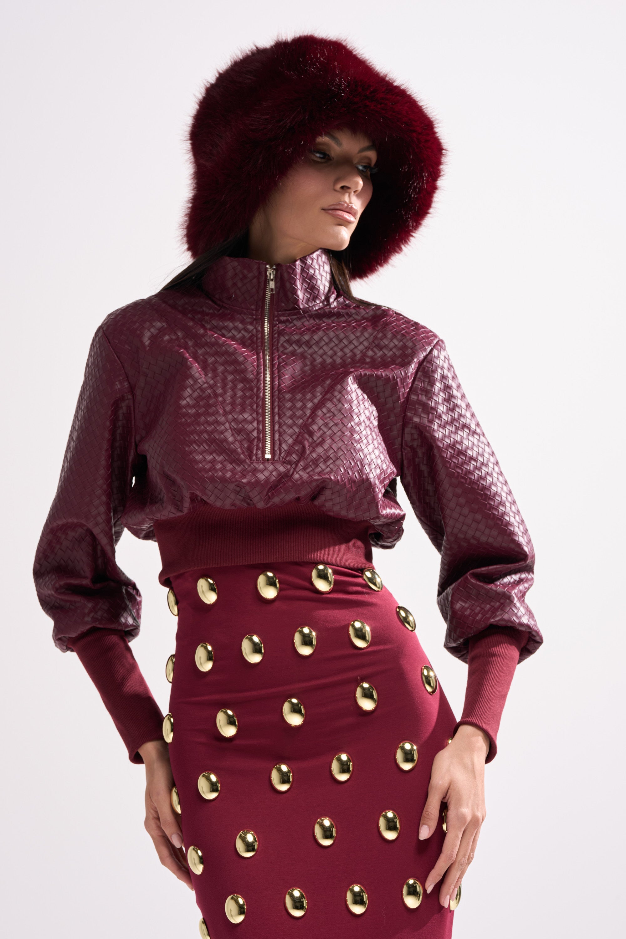 A woman wears a textured maroon jacket with a fur-trimmed hood, a matching maroon skirt with gold studs, and layers it over the VENETTA FAUX LEATHER QUARTER ZIP SWEATSHIRT, all set against a plain light background for contemporary flair.