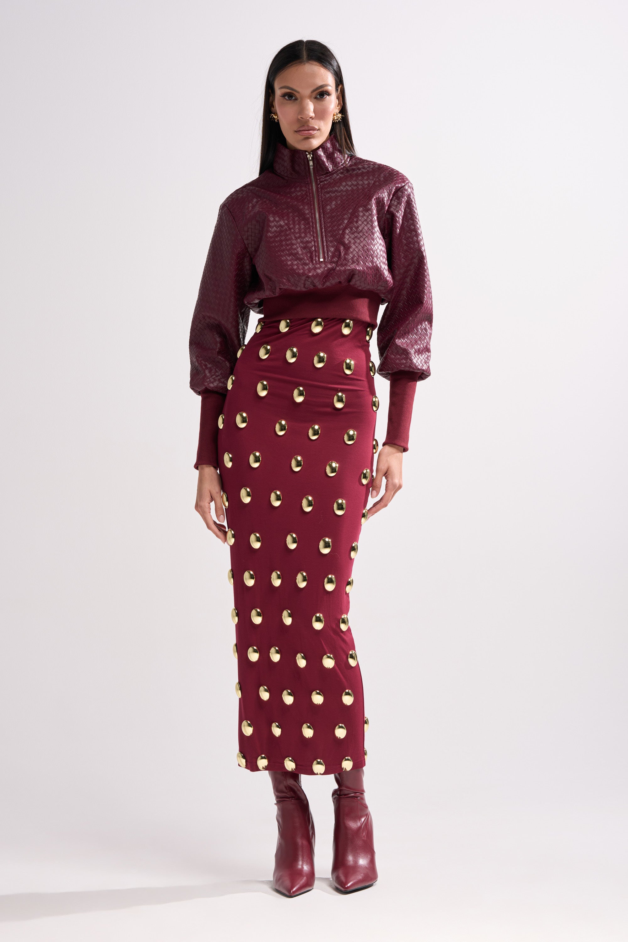 A woman poses against a plain background wearing the VENETTA FAUX LEATHER QUARTER ZIP SWEATSHIRT in maroon, paired with a matching high-waisted skirt featuring gold circular embellishments and maroon ankle boots.