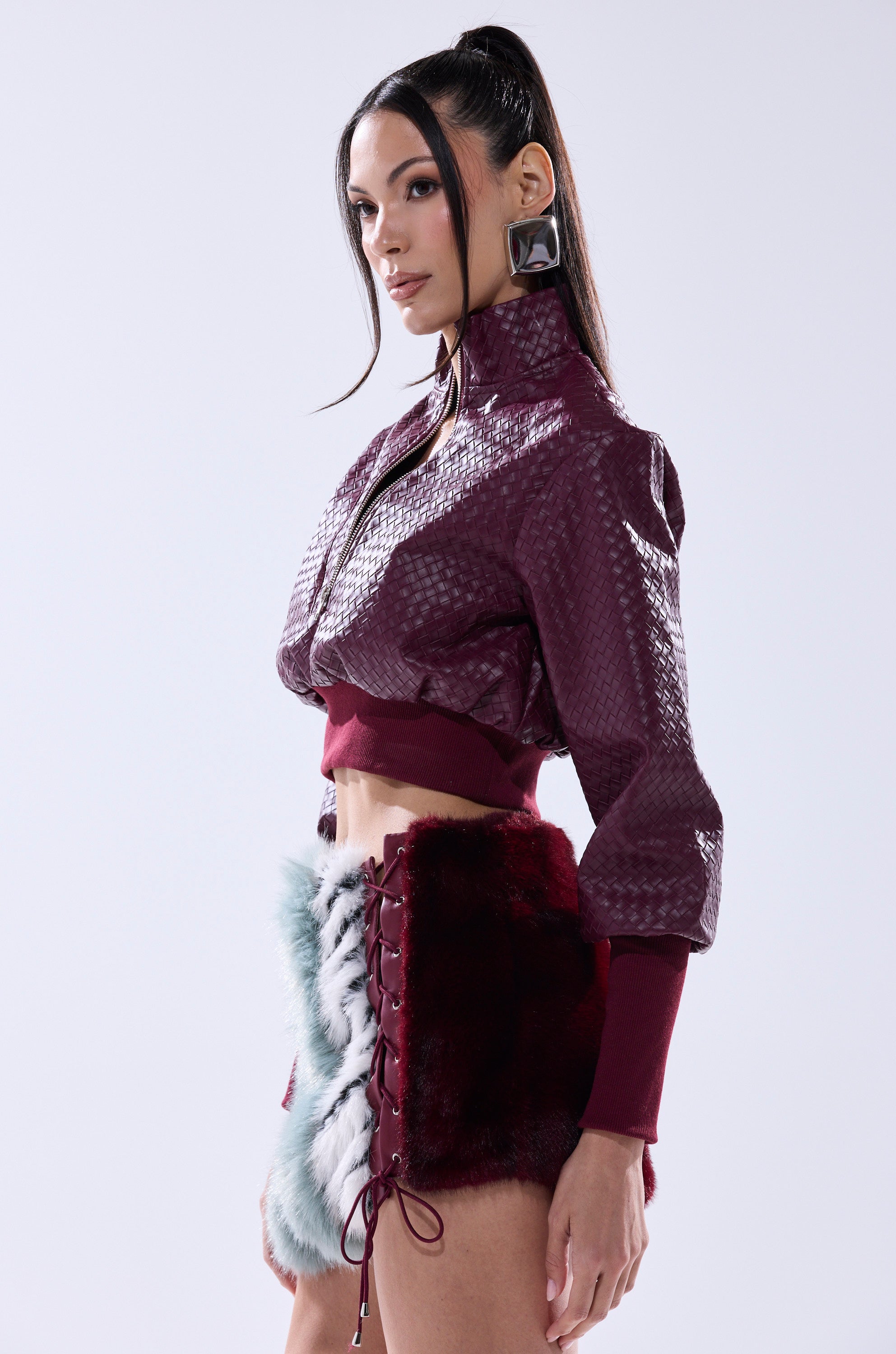 A woman poses in an AKIRA Label VENETTA Faux Leather Quarter Zip Sweatshirt, styled with large geometric earrings and a high ponytail.