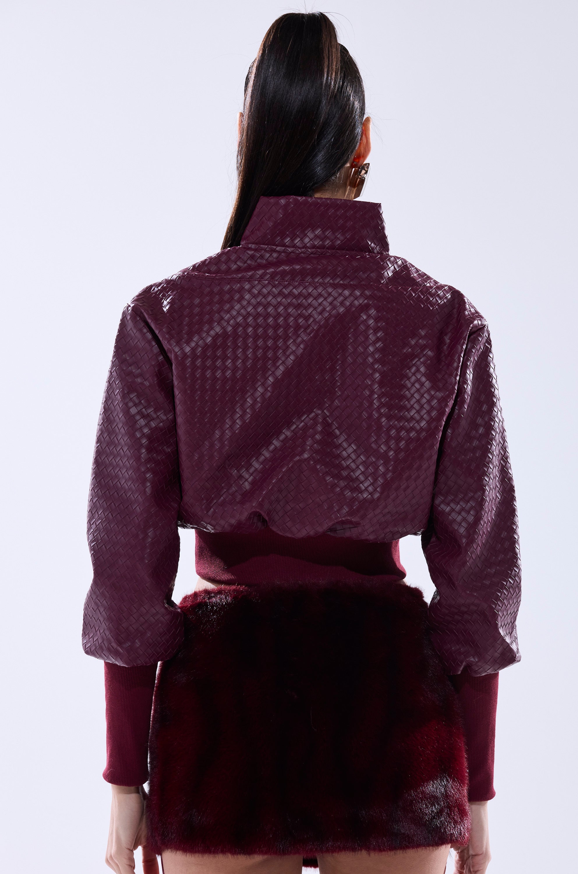 A woman with long dark hair in a ponytail is shown from behind, wearing the VENETTA FAUX LEATHER QUARTER ZIP SWEATSHIRT and a textured maroon jacket over a dark burgundy fur mini skirt, set against a plain light background.