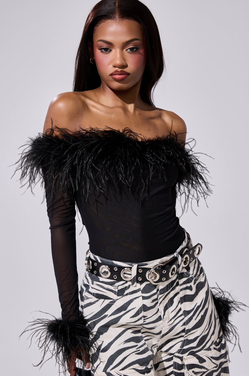 A woman poses in the FLAPPER GIRL BODYSUIT with feather details and sheer sleeves, styled with high-waisted zebra print pants and a studded belt. She has straight hair and bold makeup.