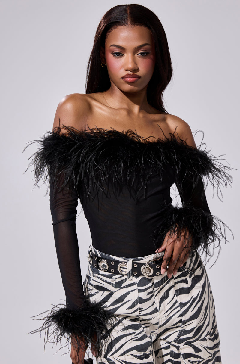 A woman with long, straight hair poses confidently against a plain light background in the FLAPPER GIRL BODYSUIT—an off-shoulder sheer mesh black top with feather detailing—paired with zebra-print pants and one hand on her hip.
