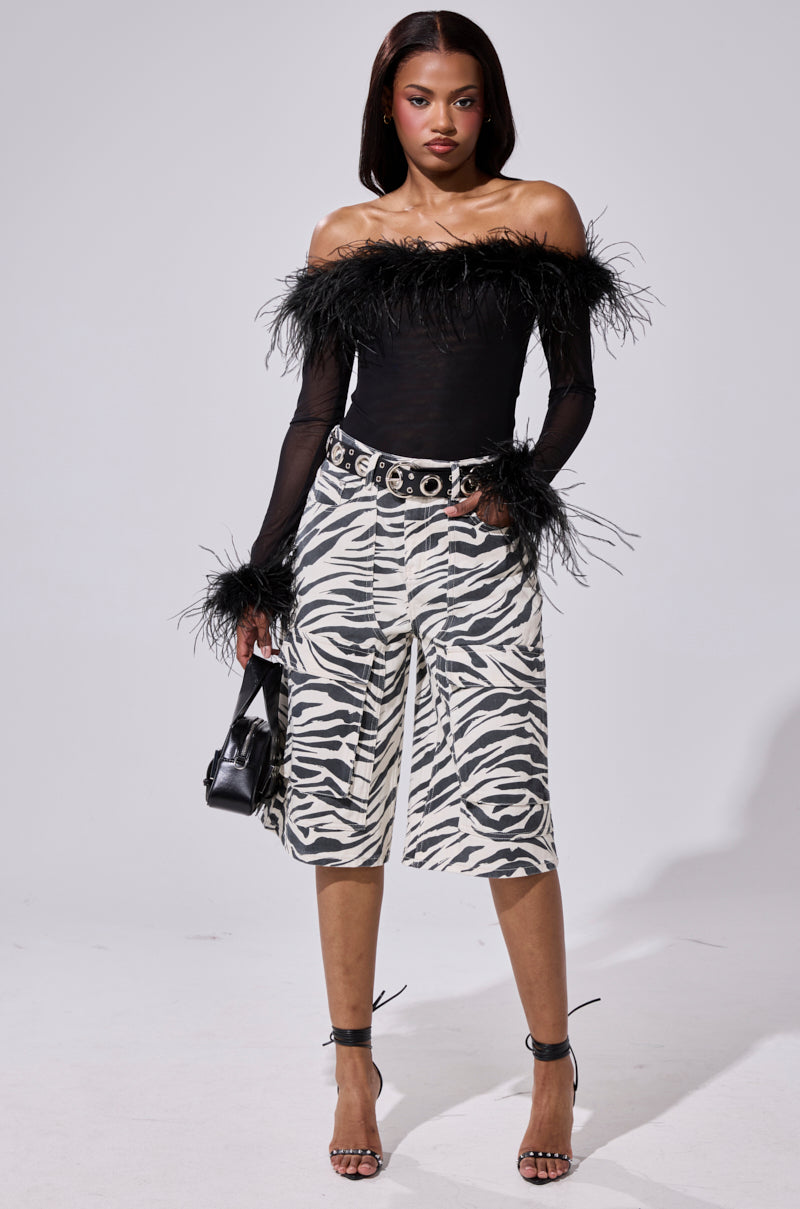 A woman exudes confidence in the FLAPPER GIRL BODYSUIT with feather details, paired with zebra-print knee-length shorts, a studded belt, black heeled sandals, and a small black handbag.