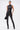 A woman stands against a white background wearing the Straight To The Point Faux Leather Legging in black with pointed heeled boots. She pairs the look with a black handbag, a wide silver cuff bracelet, and slicked-back hair.