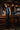 Wearing the STRAIGHT TO THE POINT FAUX LEATHER LEGGING and tall black boots, a woman poses confidently on a wooden bar counter in a dimly lit pub with rustic decor, surrounded by glasses and bar taps.