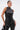 A woman with slicked-back hair confidently poses in the STRAIGHT TO THE POINT FAUX LEATHER BLOUSE, a sleeveless top with an asymmetrical hem, paired with matching black faux leather pants against a plain white background.