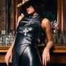 A woman in the STRAIGHT TO THE POINT FAUX LEATHER BLOUSE and a leopard print hat leans against a wooden bar, layered cross necklaces accenting her look; her face is partly hidden by the hat, with glasses filling the background.