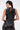 A woman with long dark hair in a ponytail wears the STRAIGHT TO THE POINT FAUX LEATHER BLOUSE, featuring a sleeveless design, asymmetrical hem, and back keyhole detail, paired with matching pants as she faces away from the camera.