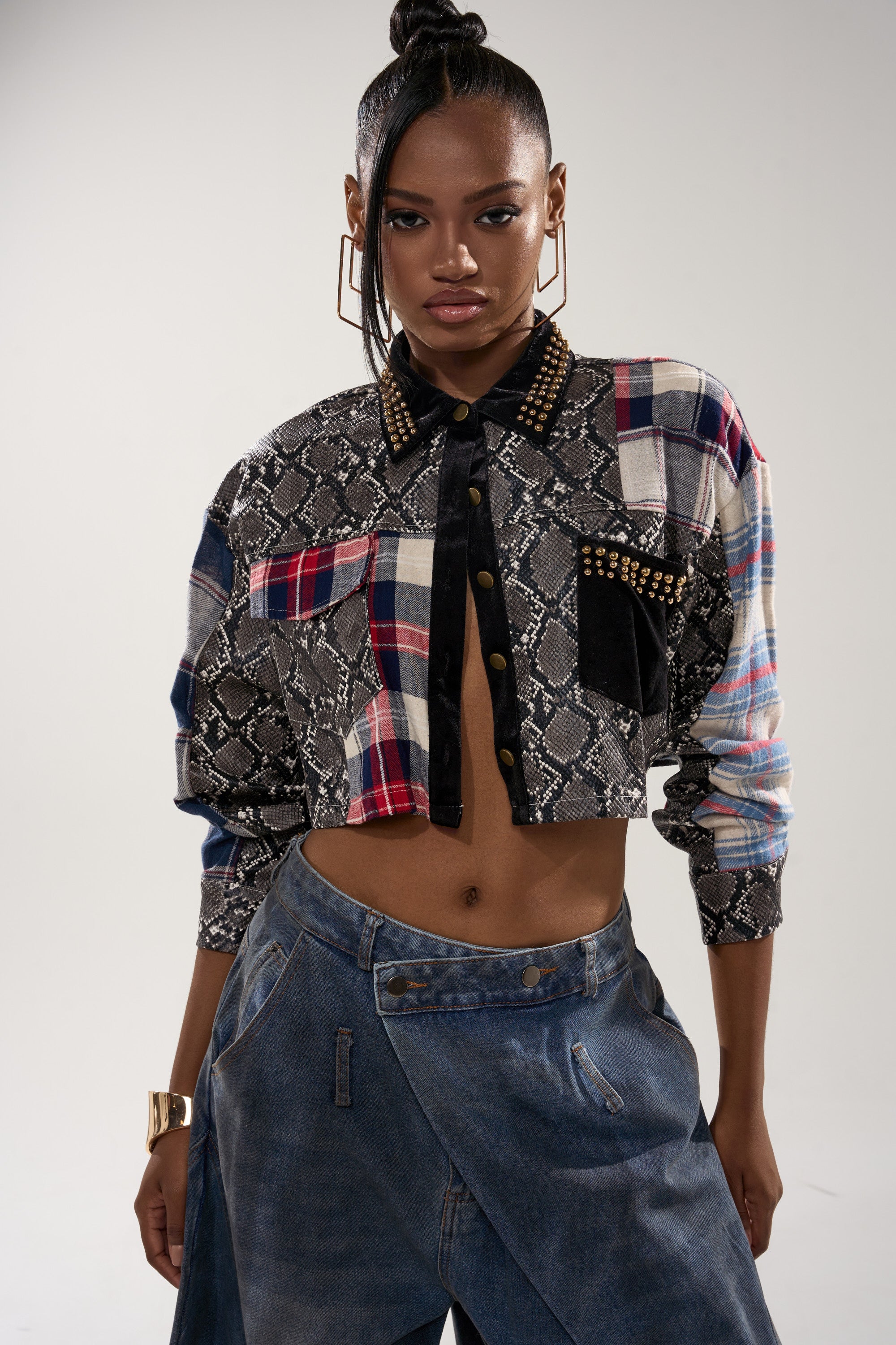 A woman exudes confidence in the GUESS WHATS NEXT PLAID BUTTON DOWN, a cropped top with snakeskin print details, styled with loose asymmetrical denim pants, hoop earrings, a high bun, and a serious expression.