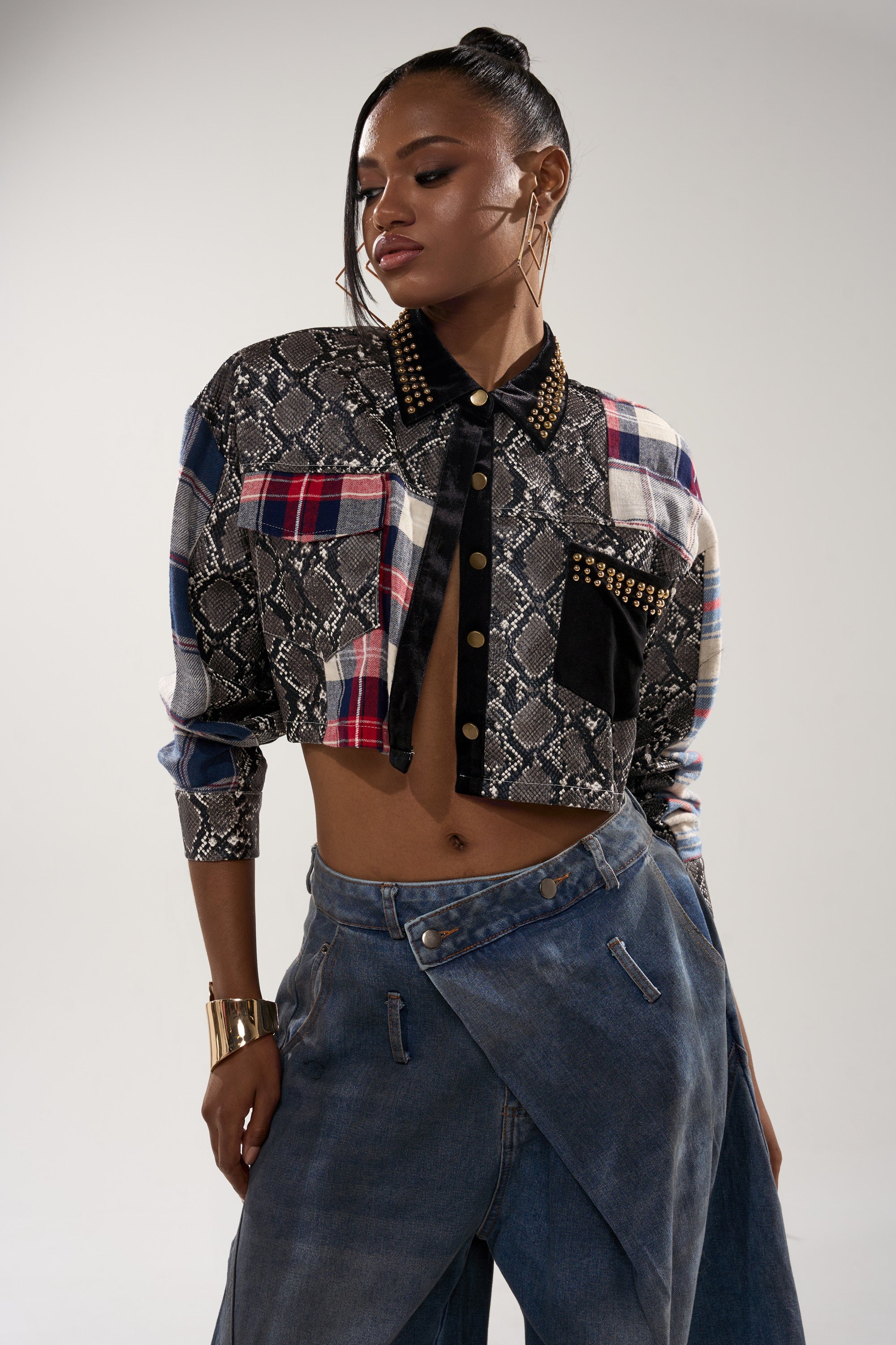 A woman poses confidently in the GUESS WHATS NEXT PLAID BUTTON DOWN, styled as a cropped fit with plaid and snakeskin print accents plus studded details, paired with high-waisted wide-leg denim and gold hoop earrings, her hair in a sleek ponytail.
