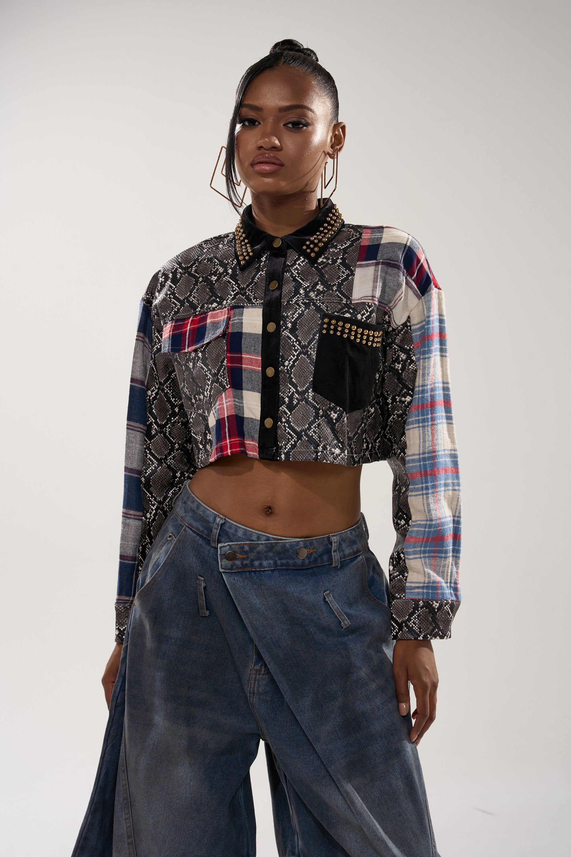 A woman stands confidently in the GUESS WHATS NEXT PLAID BUTTON DOWN, a cropped, patchwork-fit shirt with plaid, snakeskin-print, and stud details, paired with high-waisted baggy blue jeans, hoop earrings, and a high braided bun.