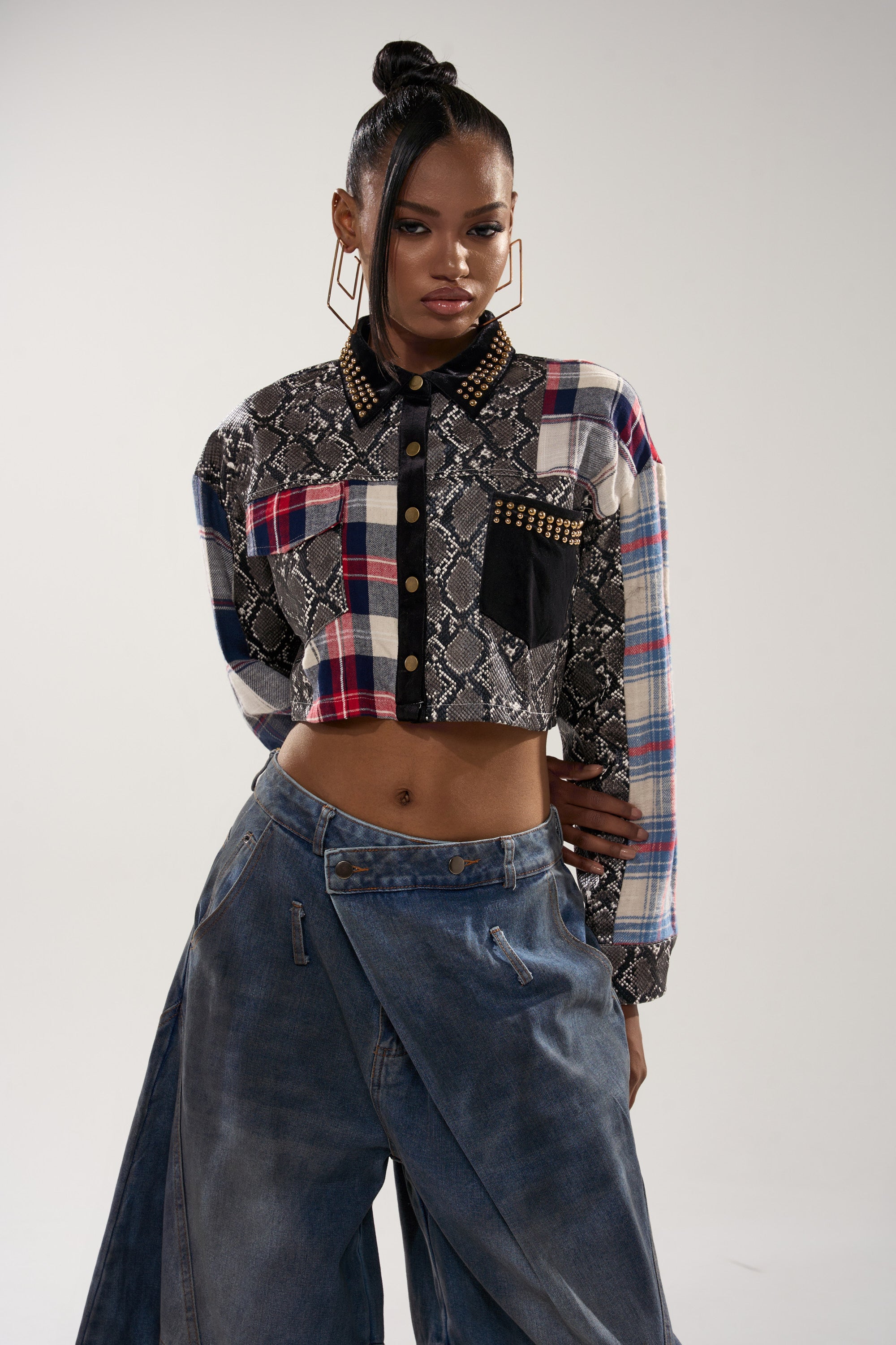 A woman stands confidently in the GUESS WHATS NEXT PLAID BUTTON DOWN—featuring snakeskin print accents and gold-studded collar—styled with wide-leg denim jeans, hoop earrings, and a high bun.