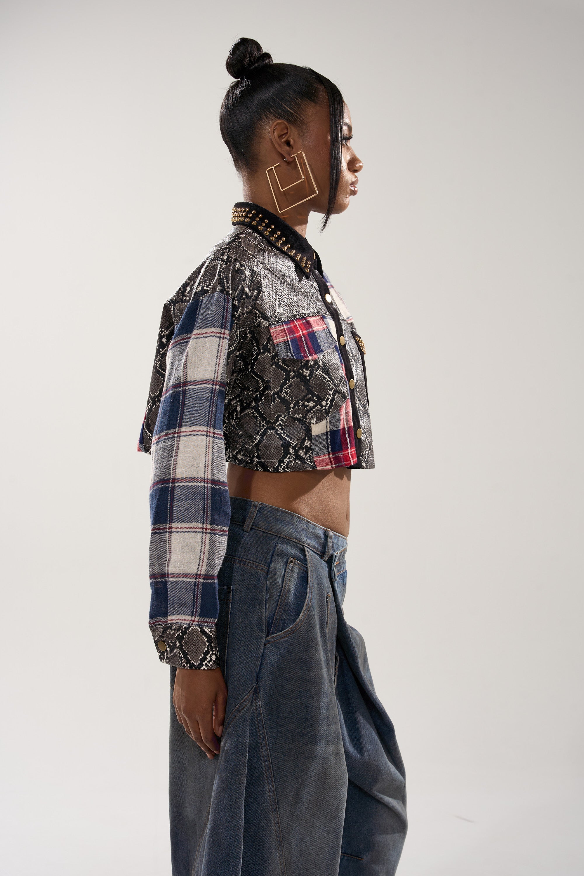 A woman in profile wears the GUESS WHATS NEXT PLAID BUTTON DOWN, styled as a cropped fit with snakeskin print accents, paired with loose denim pants and large square earrings. Her hair is in a bun, and she stands against a plain light background.