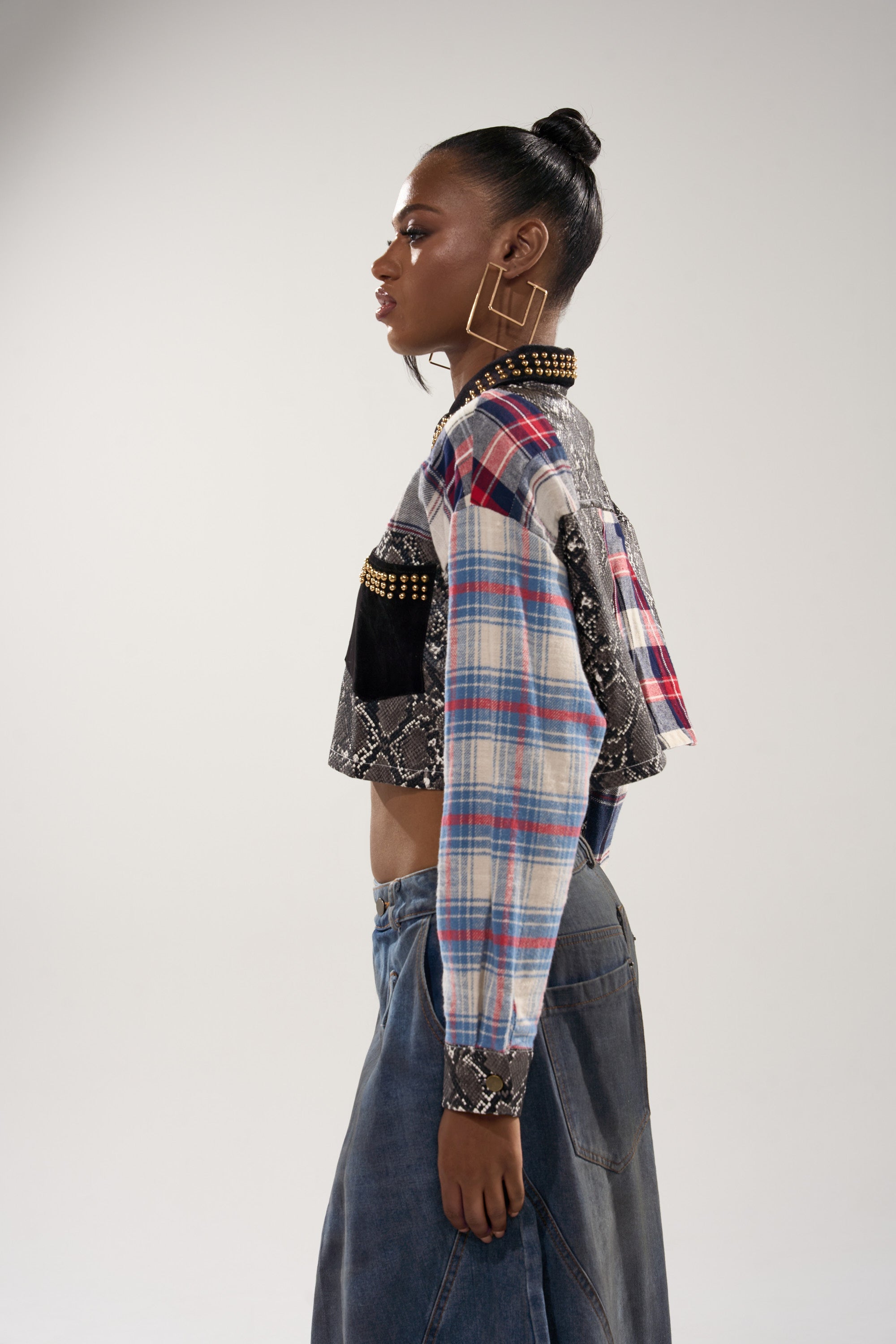 A woman in profile wears the GUESS WHATS NEXT PLAID BUTTON DOWN—a cropped fit top with plaid and snakeskin print accents and studded details—paired with wide-leg denim pants. Her hair is styled in a top bun.