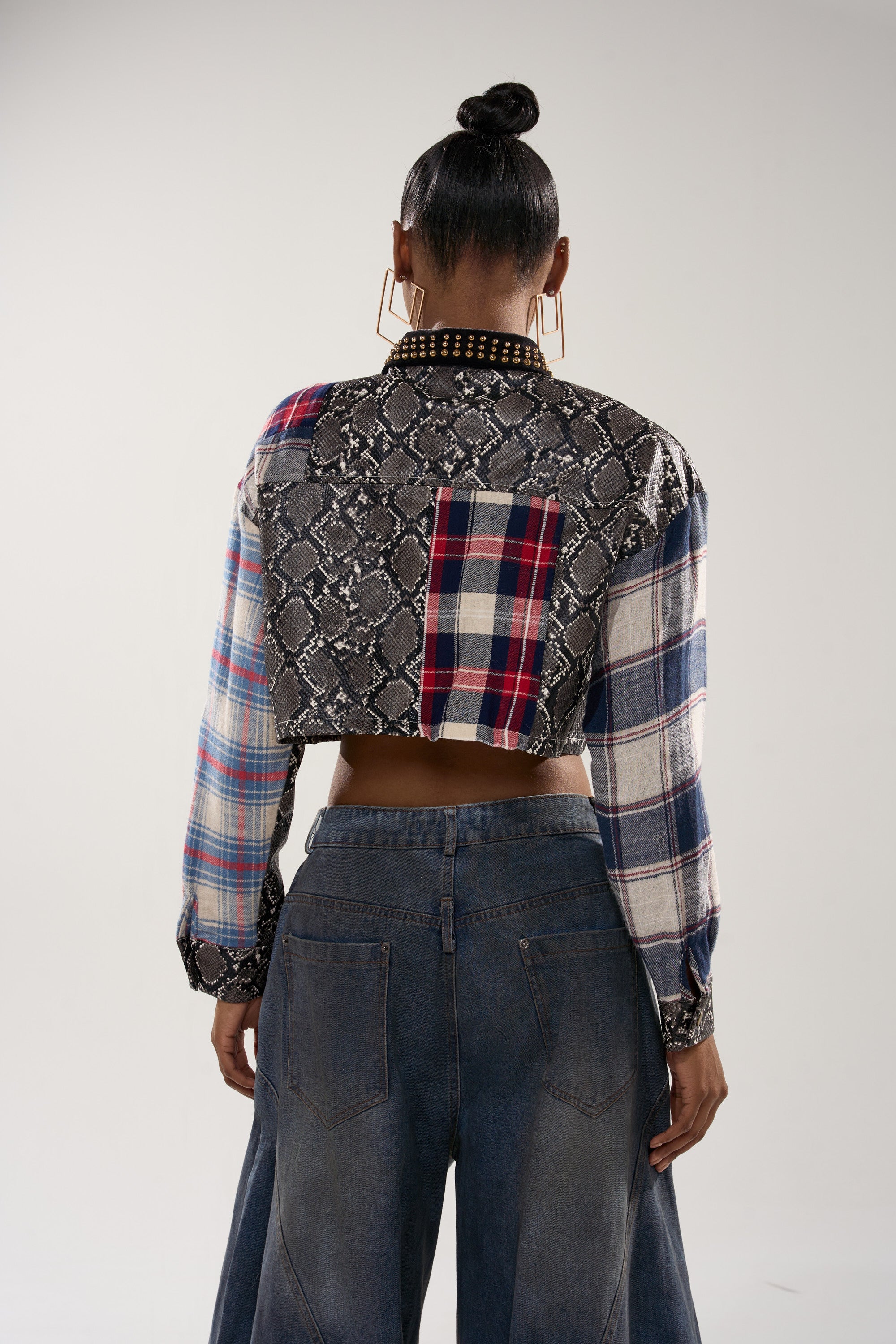 A person stands with their back to the camera, wearing wide-leg denim jeans and a cropped fit GUESS WHATS NEXT PLAID BUTTON DOWN. Their hair is in a high bun, and they have large square earrings.
