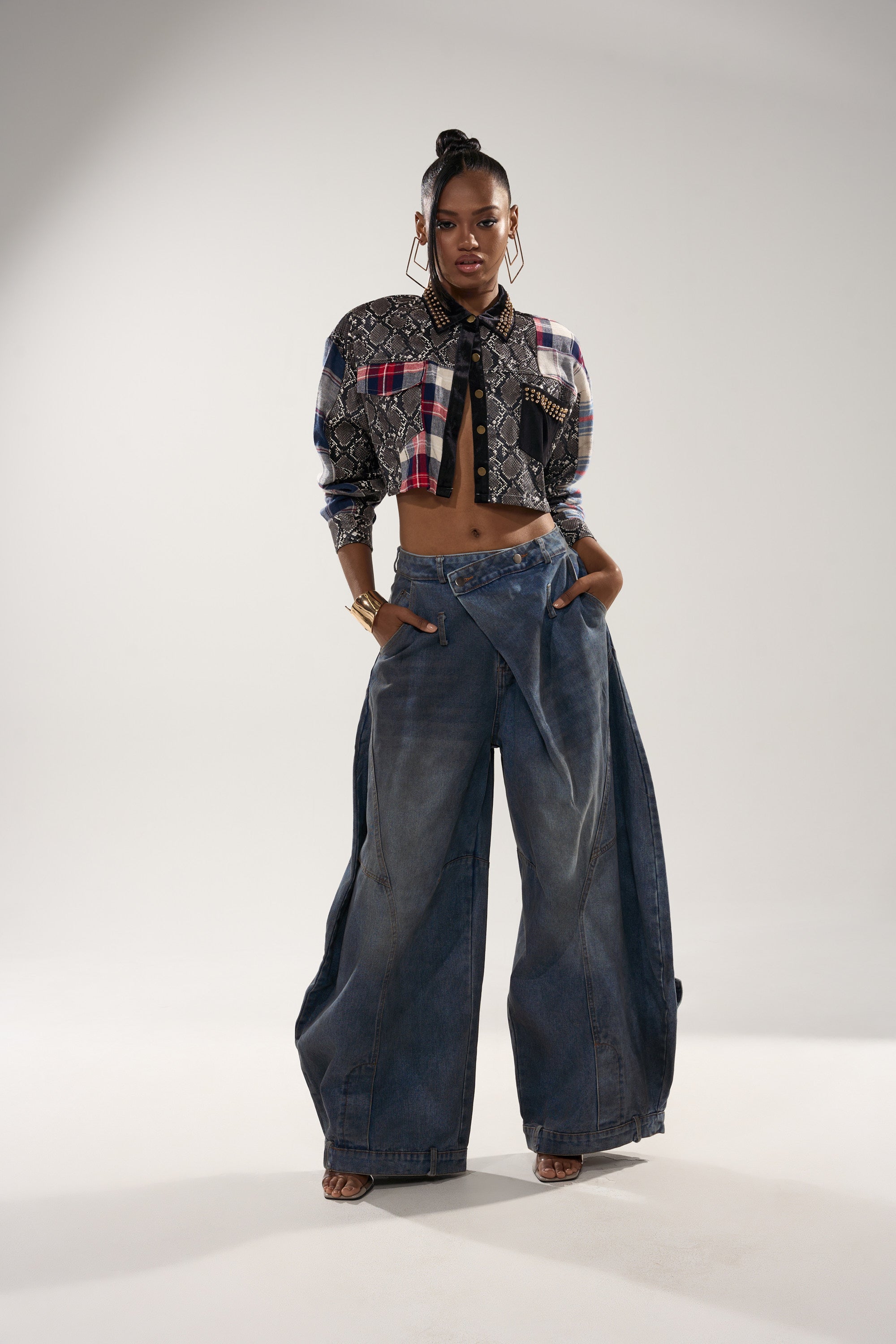 A woman stands confidently in the GUESS WHATS NEXT PLAID BUTTON DOWN, styled with high-waisted, wide-leg jeans. She accessorizes with hoop earrings, gold bracelets, and wears her hair in a high bun, hands tucked in her pockets.