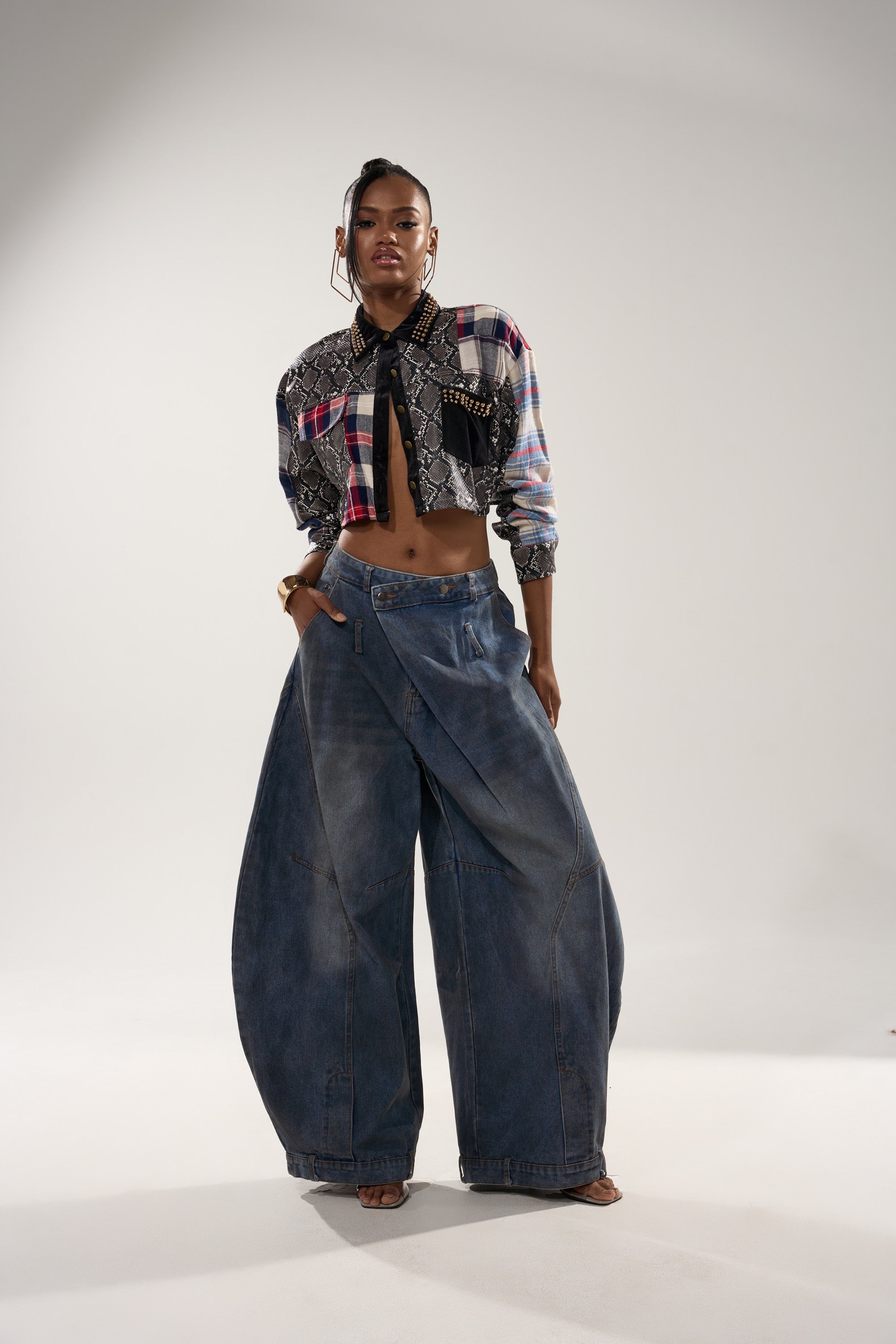 A woman poses confidently in a studio wearing the GUESS WHATS NEXT PLAID BUTTON DOWN tied at the waist with oversized baggy denim jeans, her bold and contemporary style accentuated by the light background.