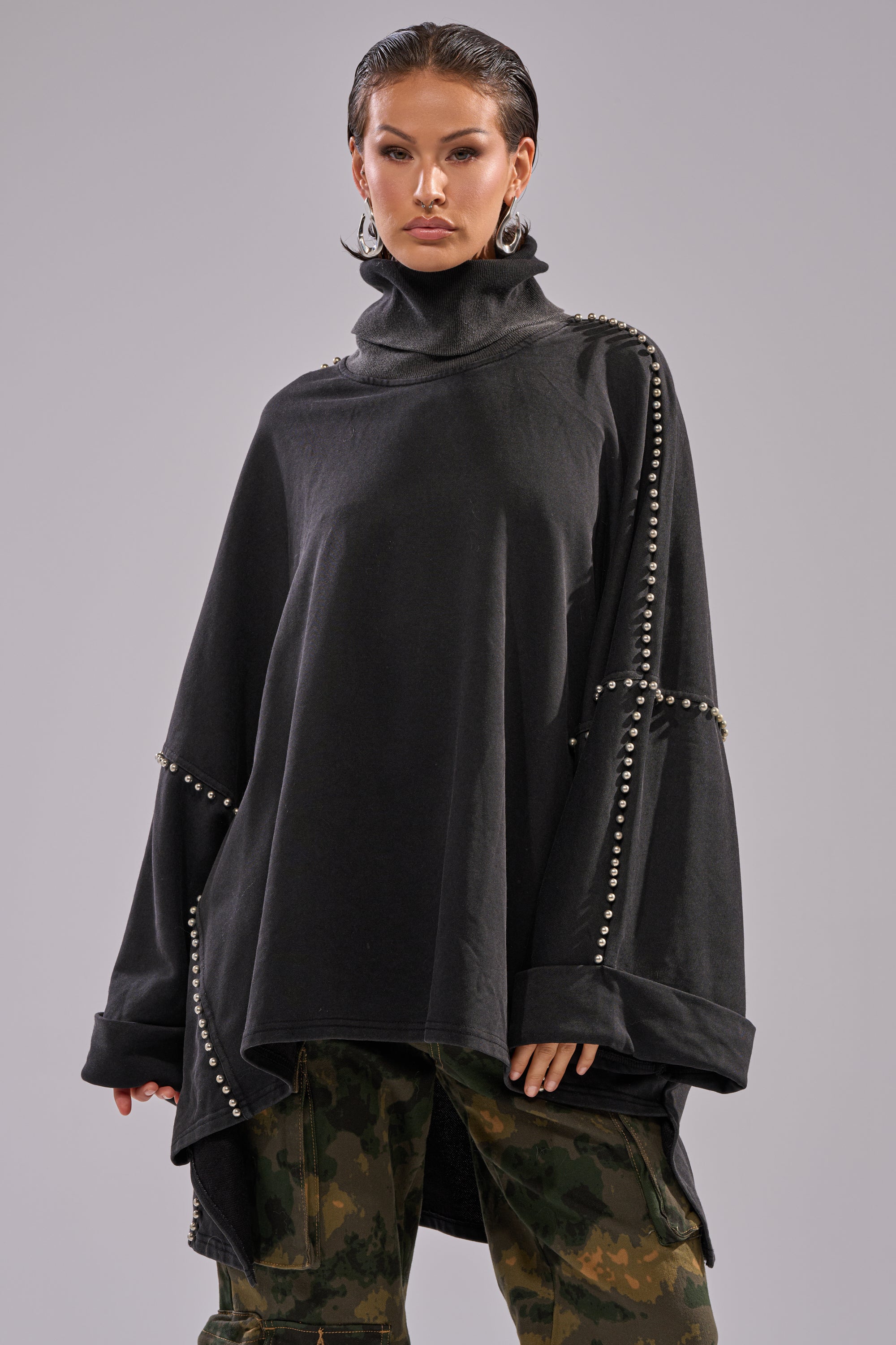 A person with slicked-back hair wears the OVER IT TURTLE NECK OVERSIZED SWEATSHIRT—silver studded with batwing sleeves—and green camouflage cargo pants, standing against a plain gray background.