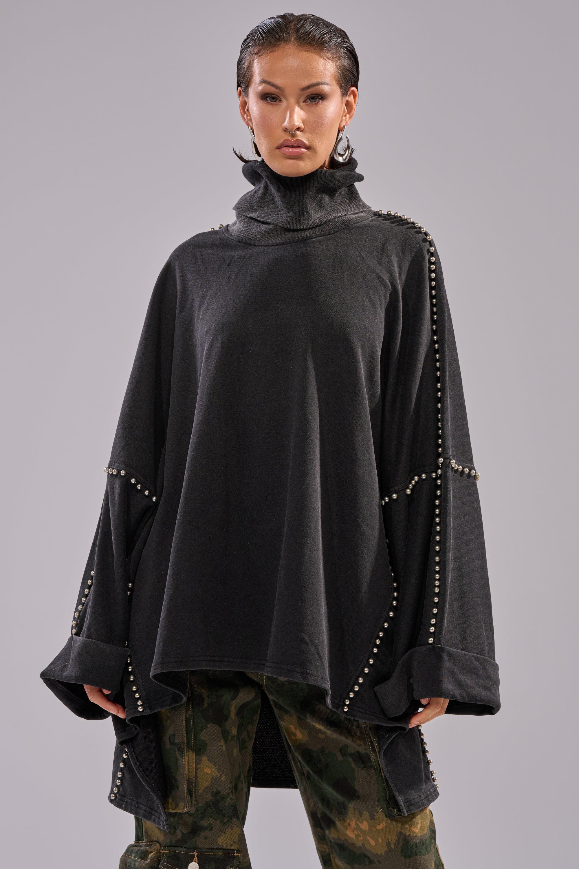 A person wears the OVER IT TURTLE NECK OVERSIZED SWEATSHIRT with batwing sleeves and metal studs over camouflage pants, standing against a plain gray background.
