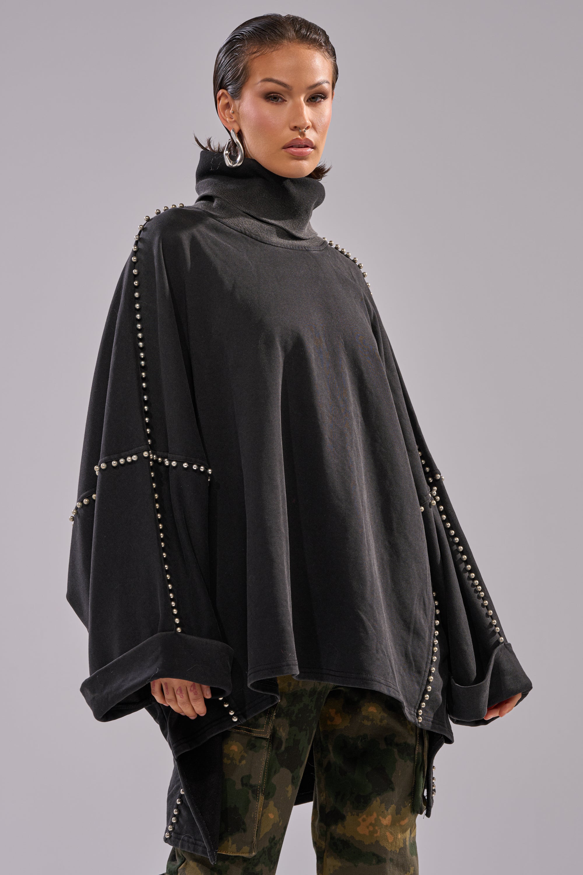 A woman stands confidently in the OVER IT TURTLE NECK OVERSIZED SWEATSHIRT with batwing sleeves and silver stud details, styled with camo pants, hoop earrings, and slicked-back hair against a plain gray background.