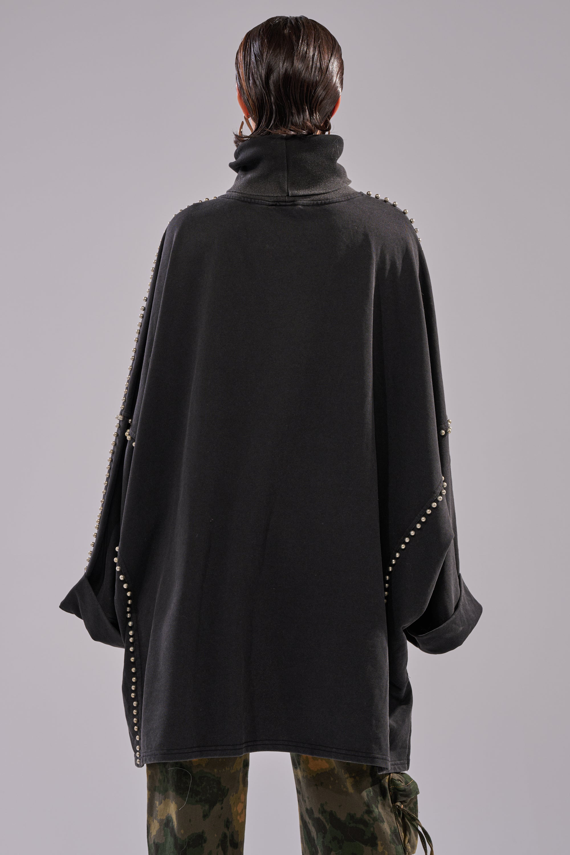 A person with wet, slicked-back hair faces away from the camera, wearing the OVER IT TURTLE NECK OVERSIZED SWEATSHIRT with metallic stud details and paired with green camouflage pants.