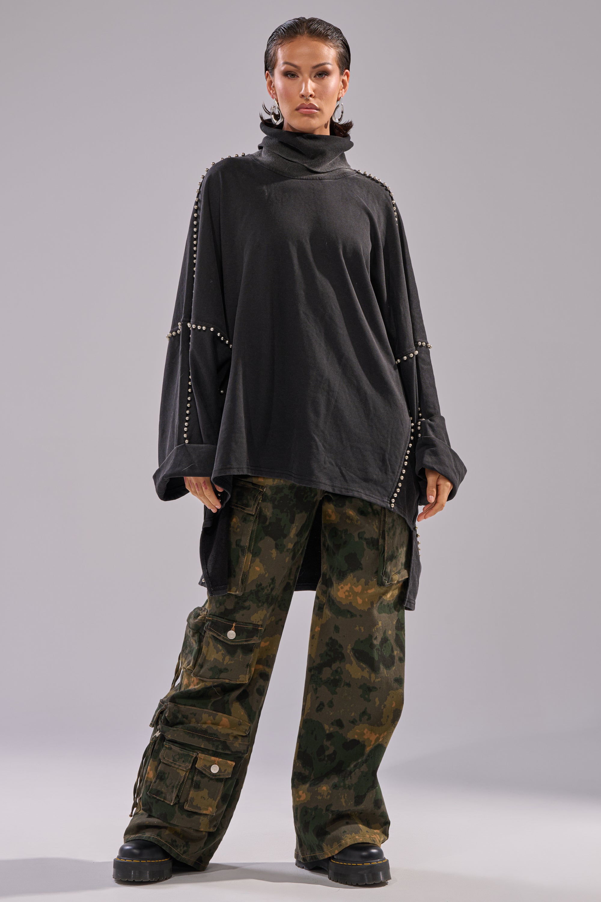A person stands confidently wearing the OVER IT TURTLE NECK OVERSIZED SWEATSHIRT with batwing sleeves and studded details, paired with loose camouflage cargo pants and chunky black shoes, against a plain light background.