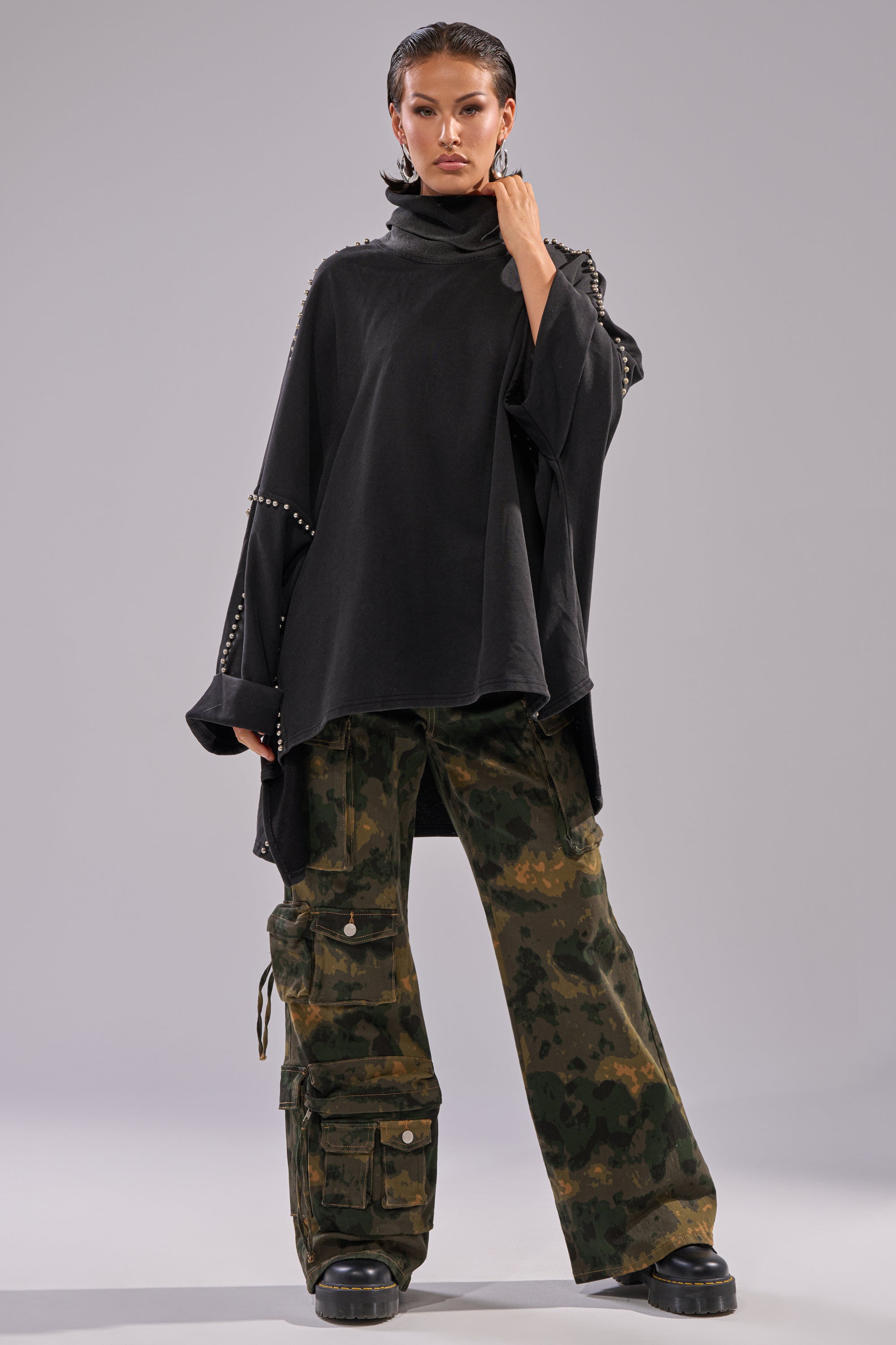 A person stands confidently in the OVER IT TURTLE NECK OVERSIZED SWEATSHIRT, styled with camouflage cargo pants and black chunky shoes against a plain gray background.