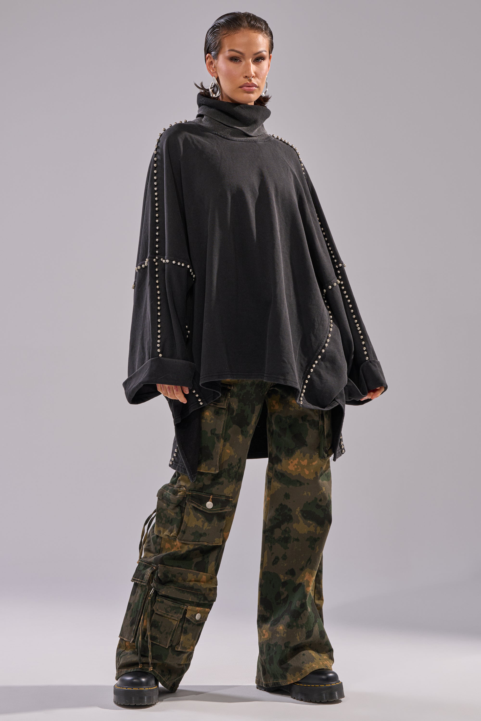 Confidently posing against a gray background, this model wears the OVER IT TURTLE NECK OVERSIZED SWEATSHIRT with batwing sleeves and metal studs, paired with oversized camouflage cargo pants and chunky black shoes.