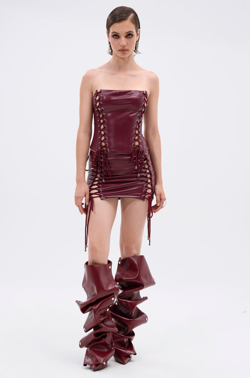 A person wears the YOU KNOW I WANT YOU BABY LACE UP MINI SKIRT in burgundy faux leather with lace-up sides, styled with oversized slouchy thigh-high boots, standing against a plain light background. 3