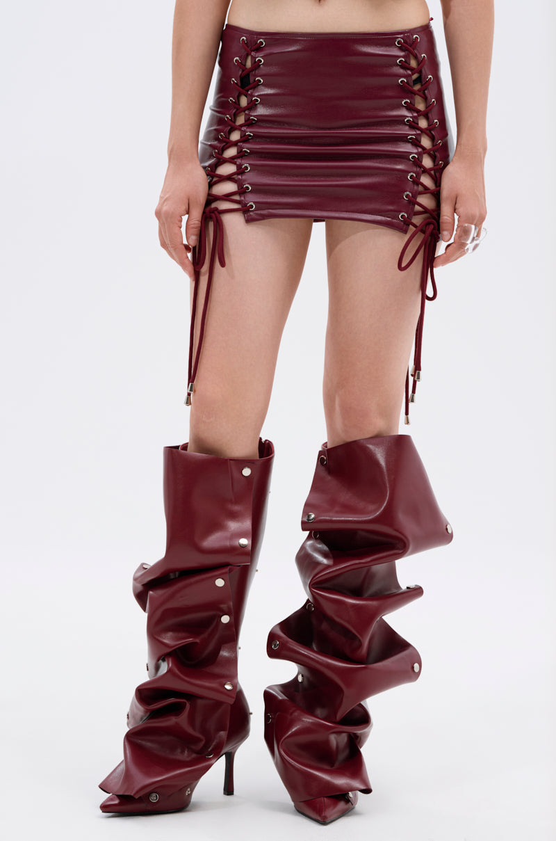 Someone models the YOU KNOW I WANT YOU BABY LACE UP MINI SKIRT paired with tall, slouchy high-heeled boots with snap buttons, standing against a plain white background. 0